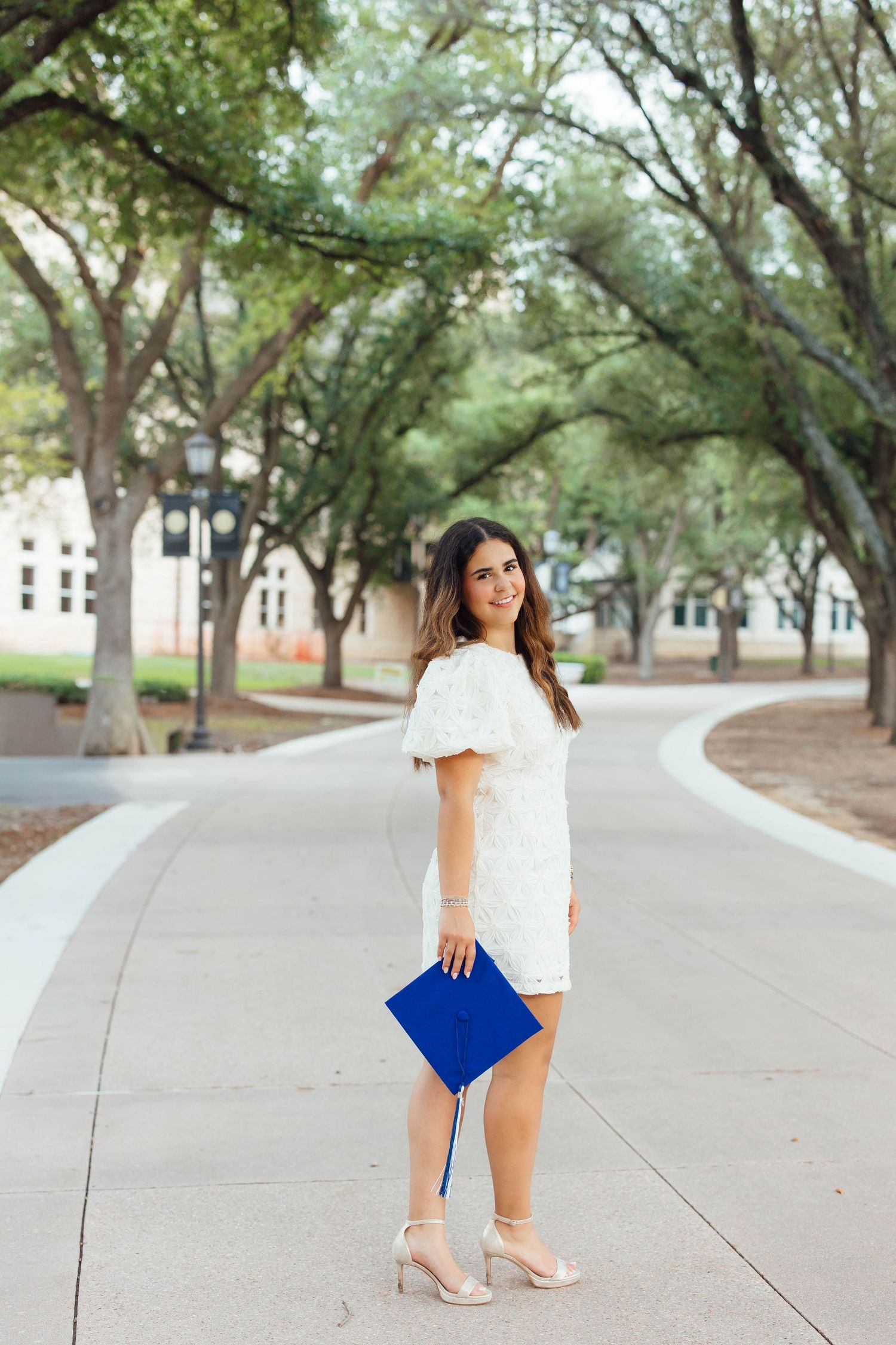 Brunette woman in white dress holding blue graduation cap on tree-lined campus walkway.