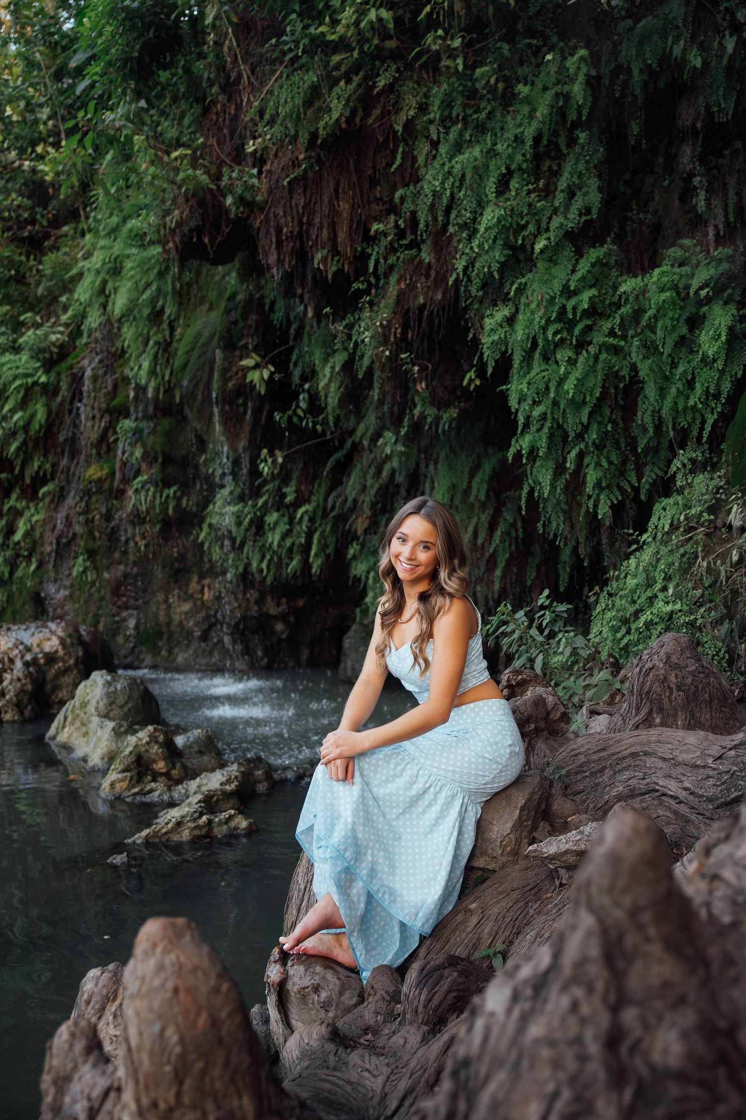 Smiling young woman in light blue sundress sitting on rocks by a lush tropical waterfall.