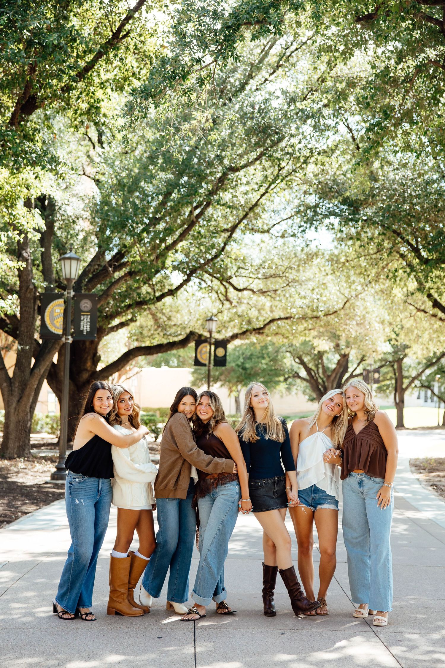 Seven young women in casual outfits pose together on a tree-lined campus walkway.