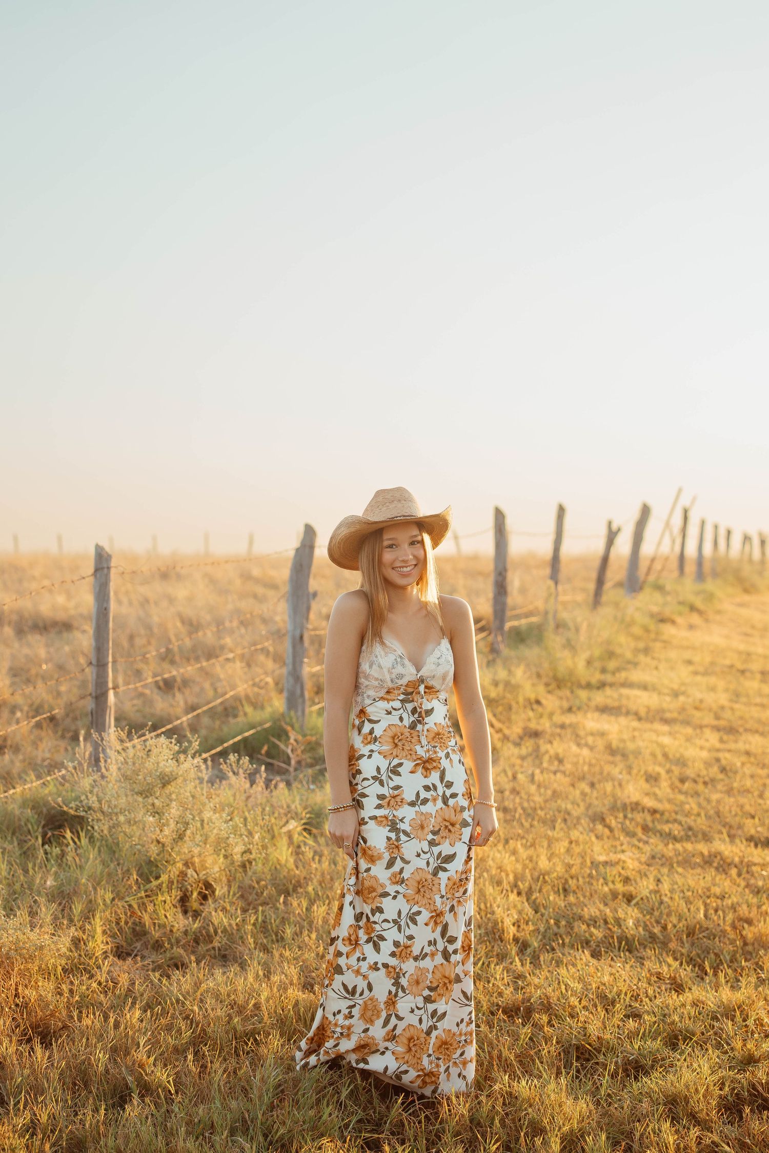 A smiling blonde woman in a floral maxi dress and straw cowboy hat stands by a fence at golden hour.