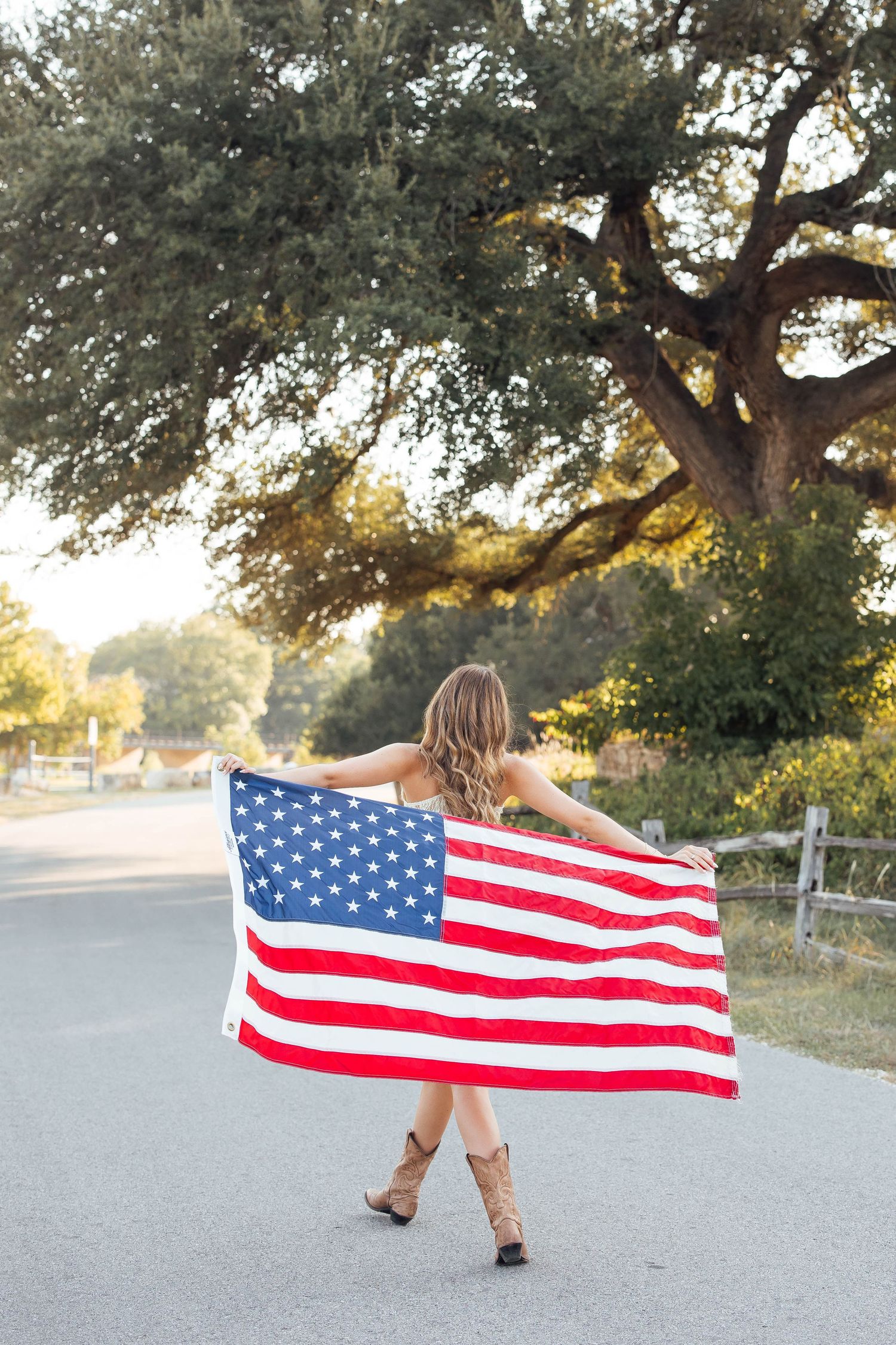 Young woman in cowboy boots holding American flag on a tree-lined road at golden hour.