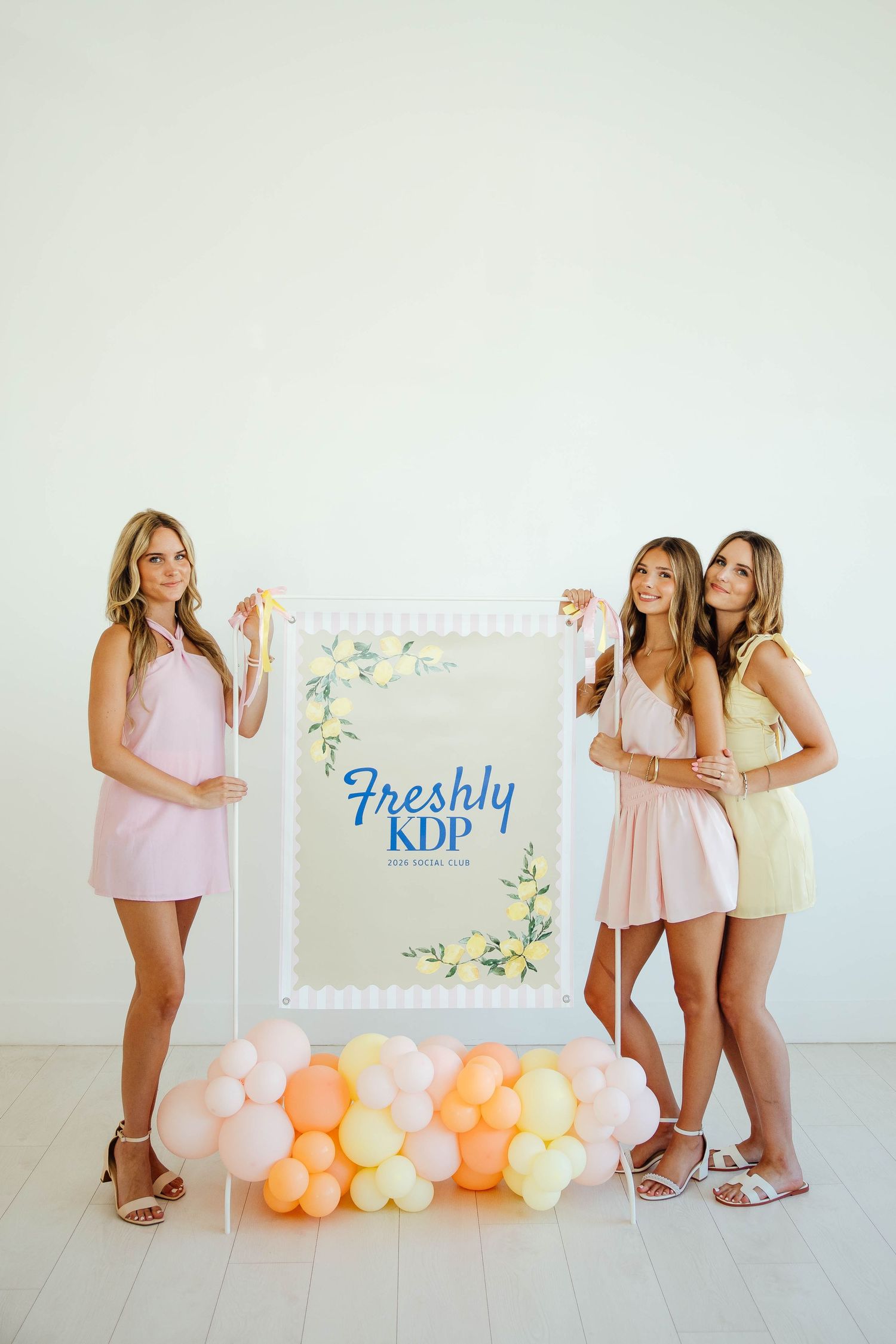 Three women in pastel dresses pose with a Freshly KDP social club sign surrounded by colorful balloons.