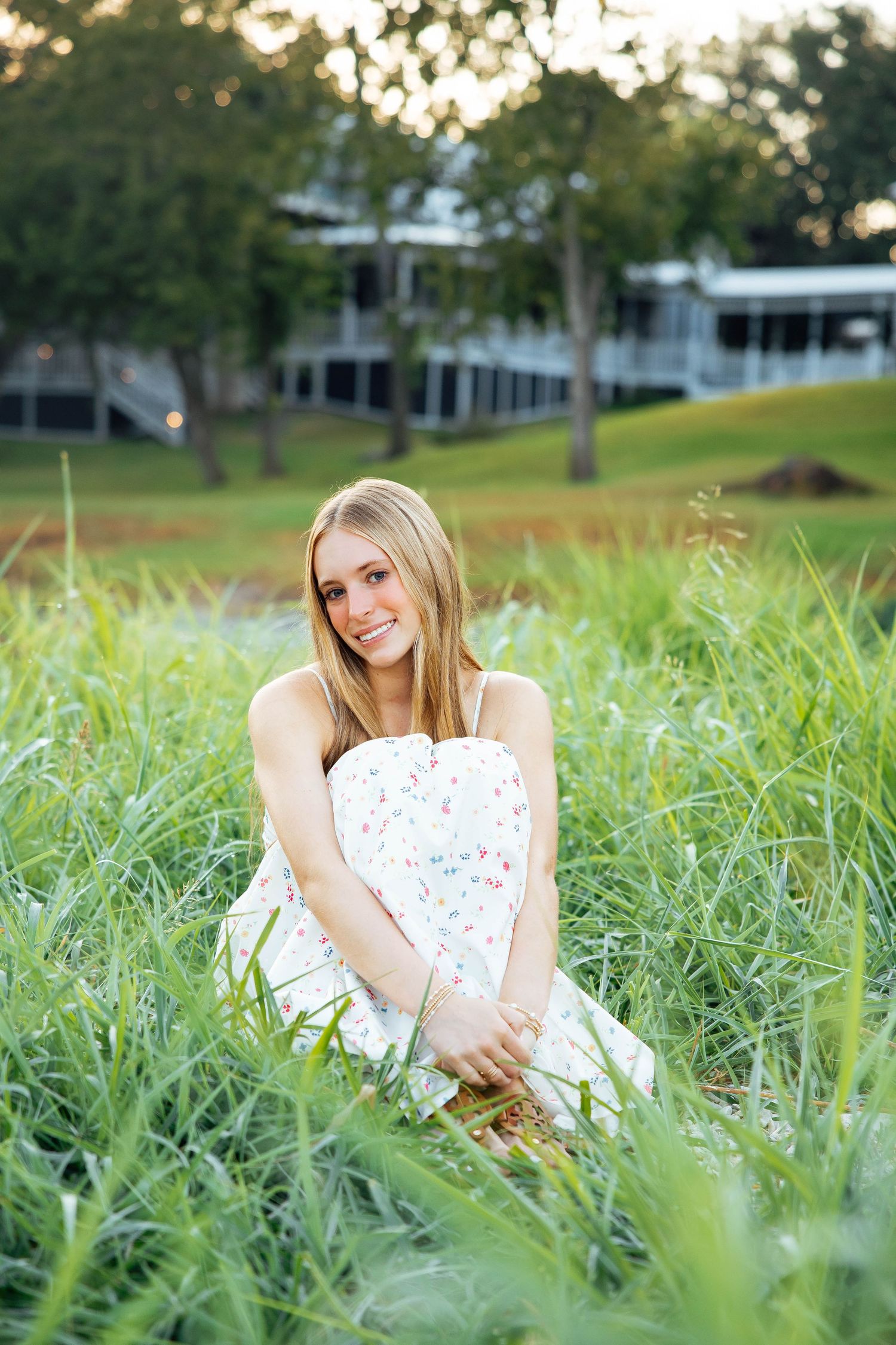 Smiling blonde teen in white floral sundress sits in tall green grass for senior portrait.