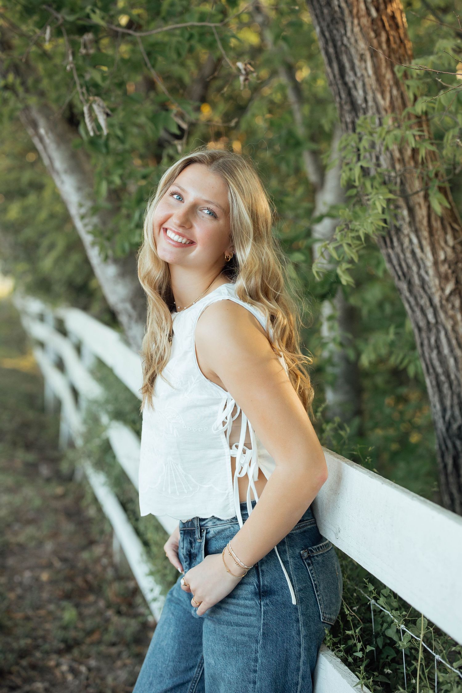 A smiling blonde teen in a white crop top and jeans leans against a white fence among trees.