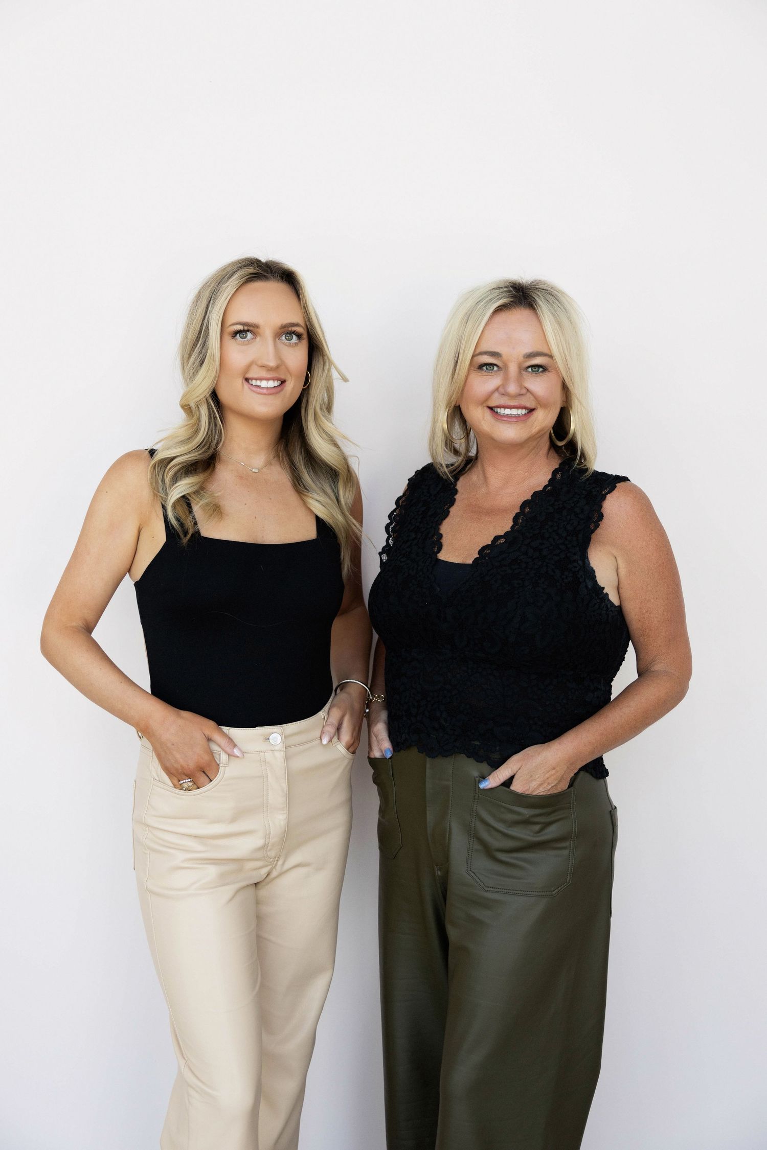 Two blonde women in black tops smiling together, one in beige pants and one in olive green trousers, against white background.