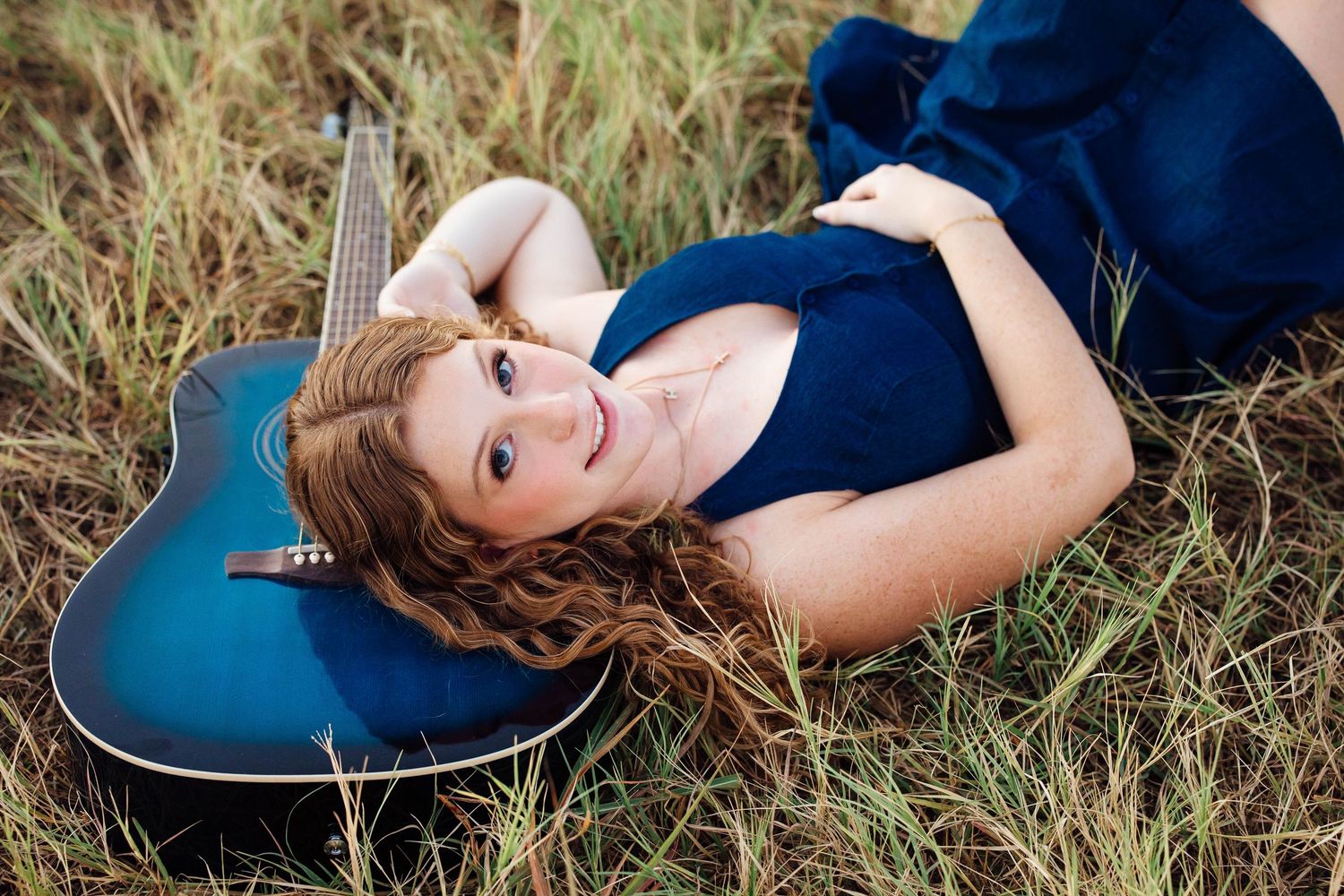 Young woman in blue dress lying in grass field with acoustic guitar outdoors.