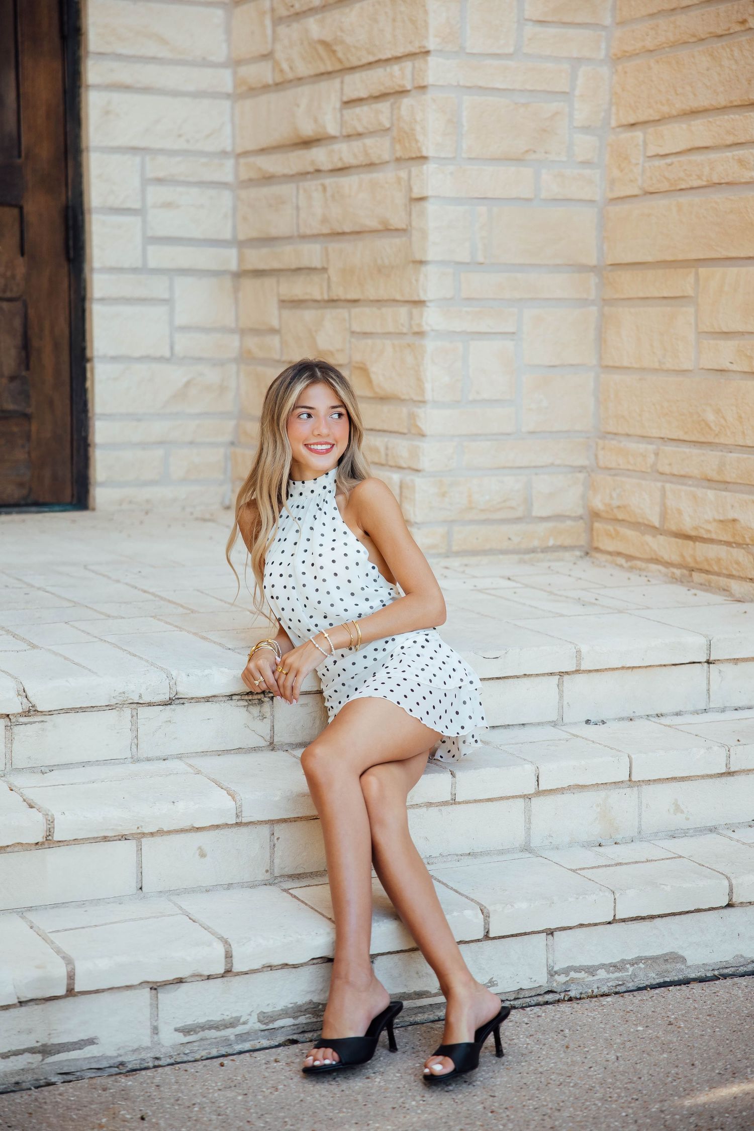 Blonde woman in white polka dot halter dress sits smiling on stone steps beside brick building.