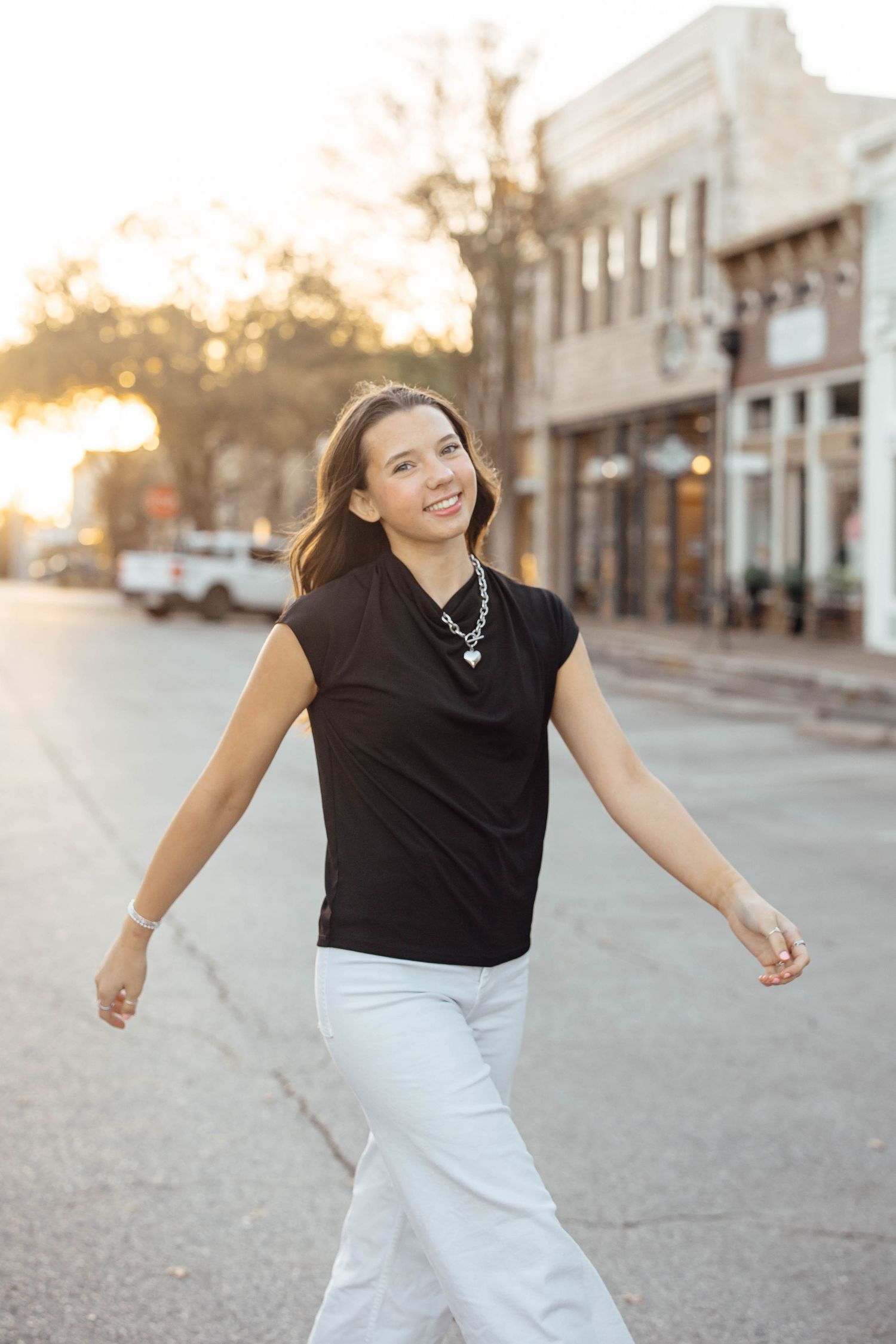 Smiling young woman in black top and white pants walking on a small town street at sunset.