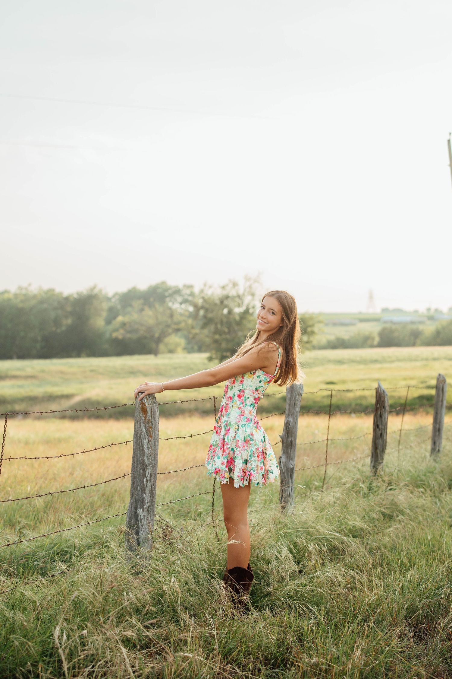 Smiling girl in floral sundress and boots poses by barbed wire fence in golden countryside field.