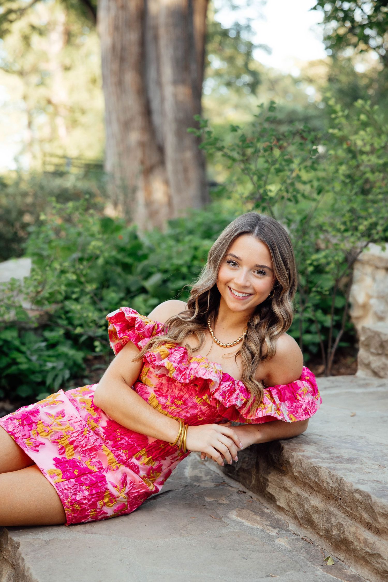 Smiling brunette woman in pink floral off-shoulder dress posing on stone steps outdoors.