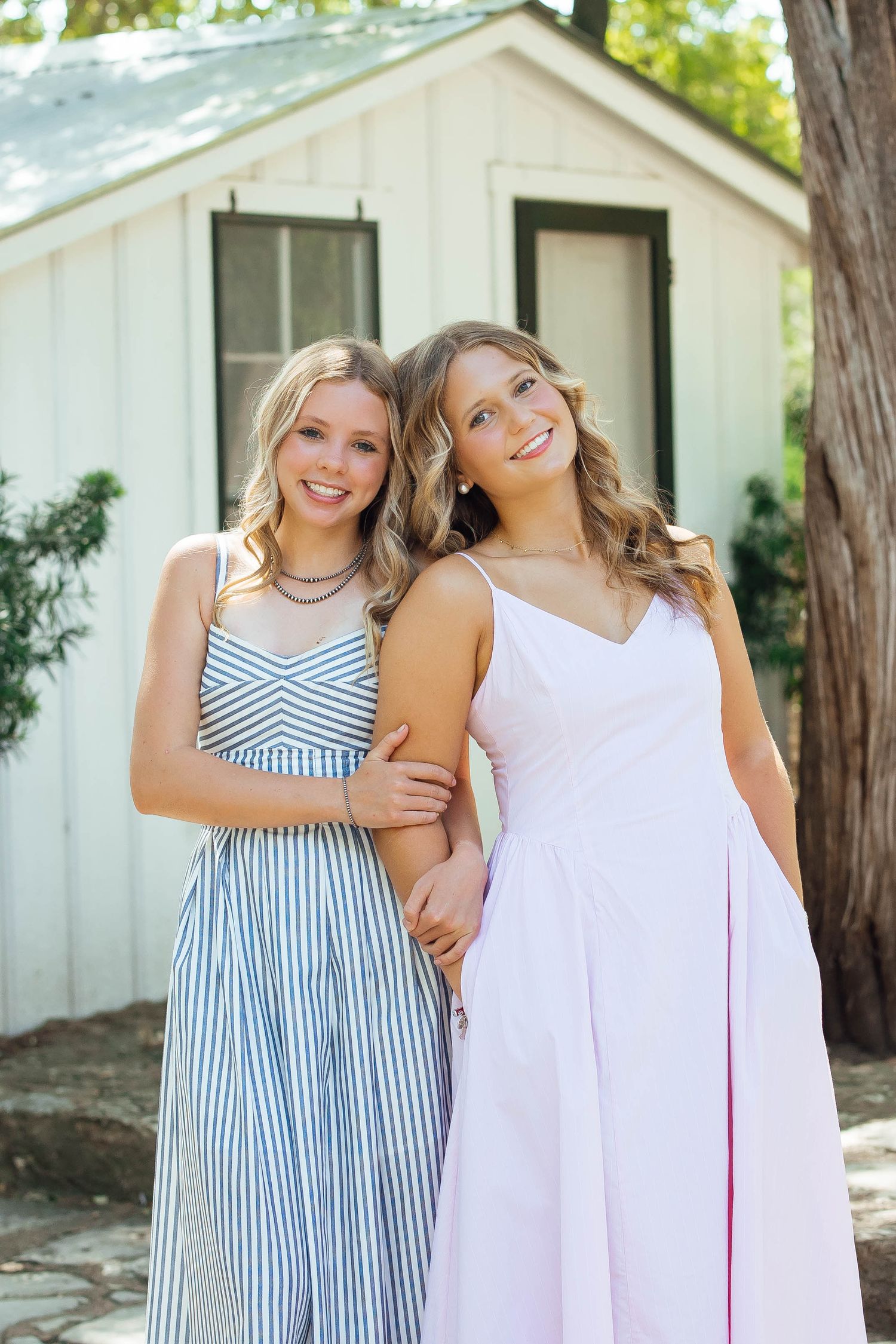 Two smiling young women in formal dresses pose together outside a white cottage.