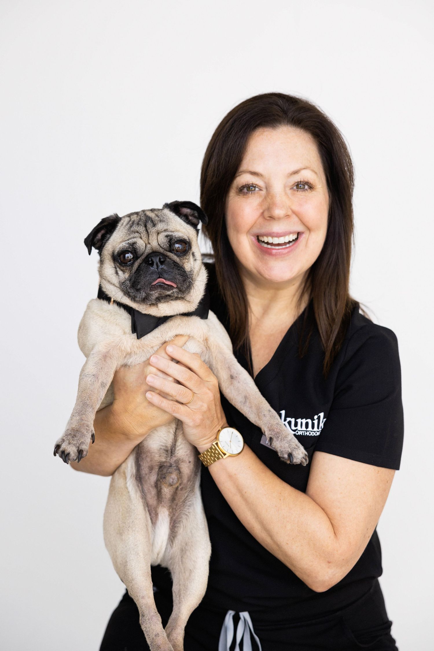 Smiling brunette woman in black scrubs holding an adorable fawn pug dog against a white background.