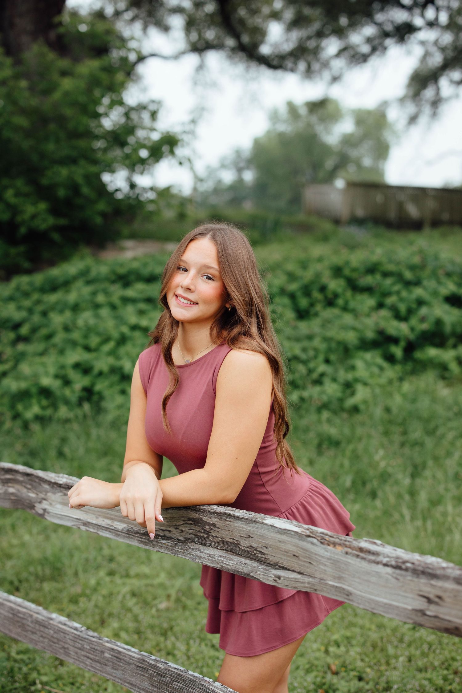 Smiling teenage girl in mauve dress leans on rustic wooden fence in lush green outdoor setting.