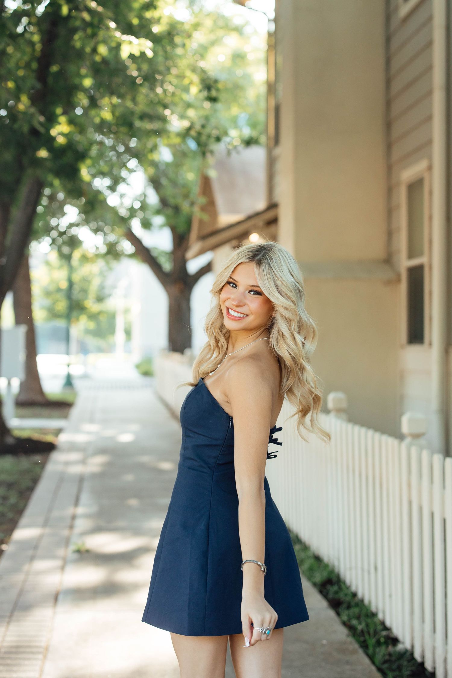 Smiling blonde woman in navy blue mini dress walks along tree-lined sidewalk near white picket fence.