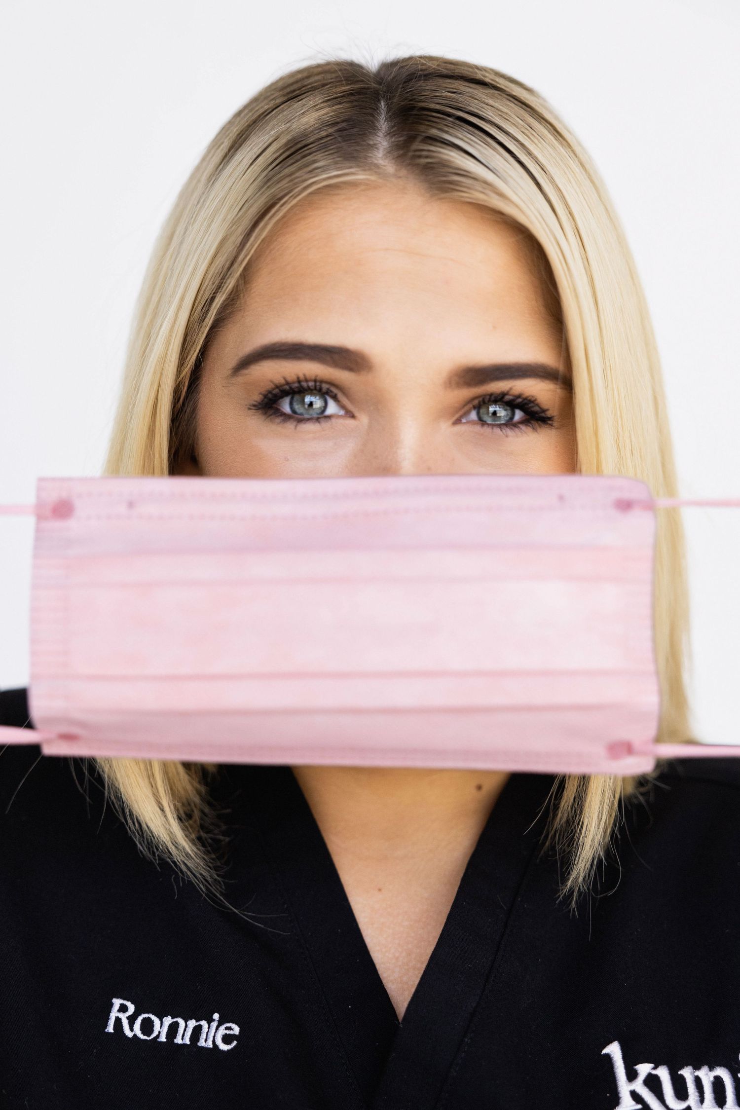 Blonde woman in black scrubs holding a pink disposable face mask in front of her mouth with blue eyes visible.