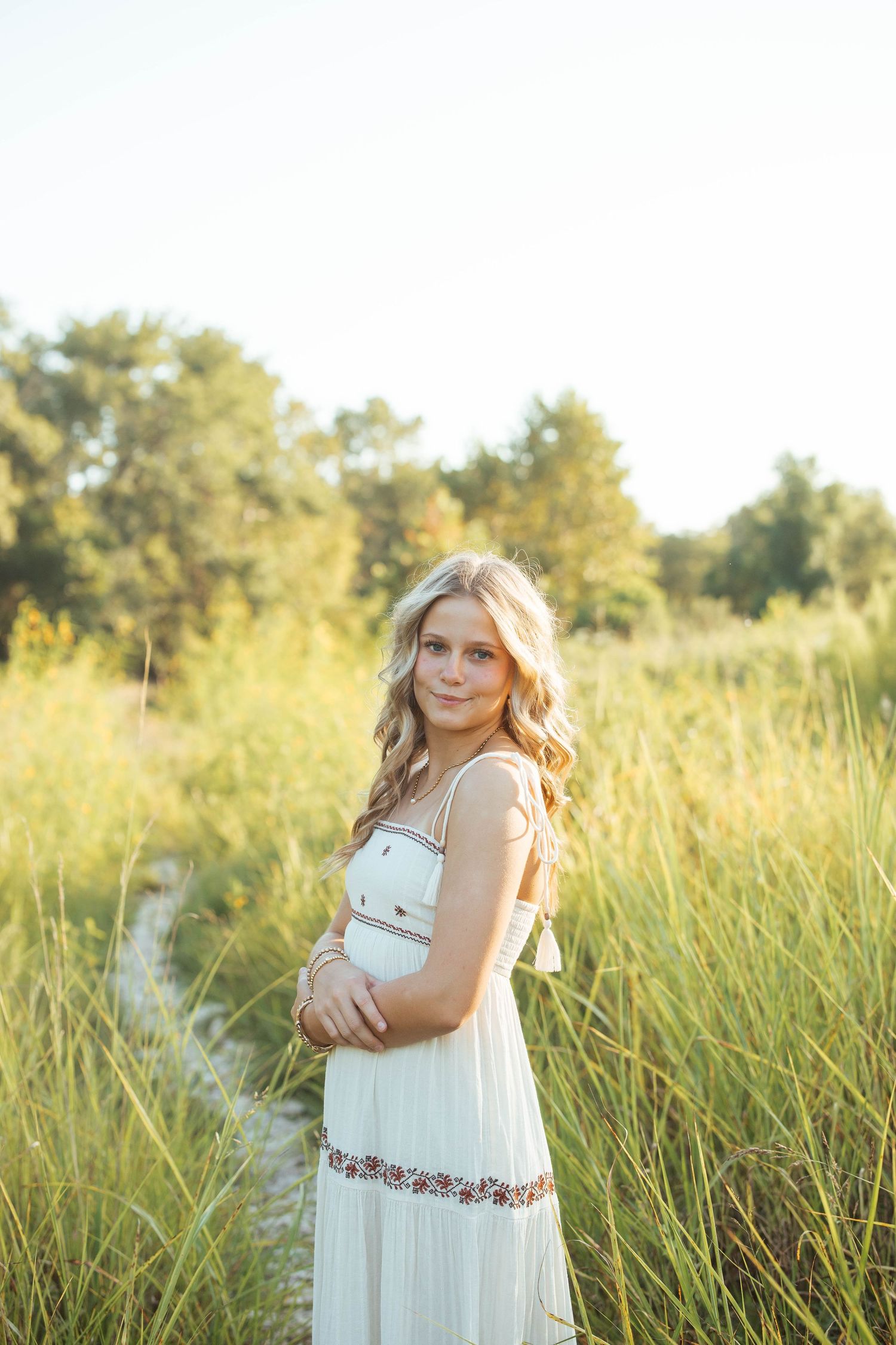 Blonde teenage girl in white maxi dress standing in tall golden grass during a sunny outdoor photo session.