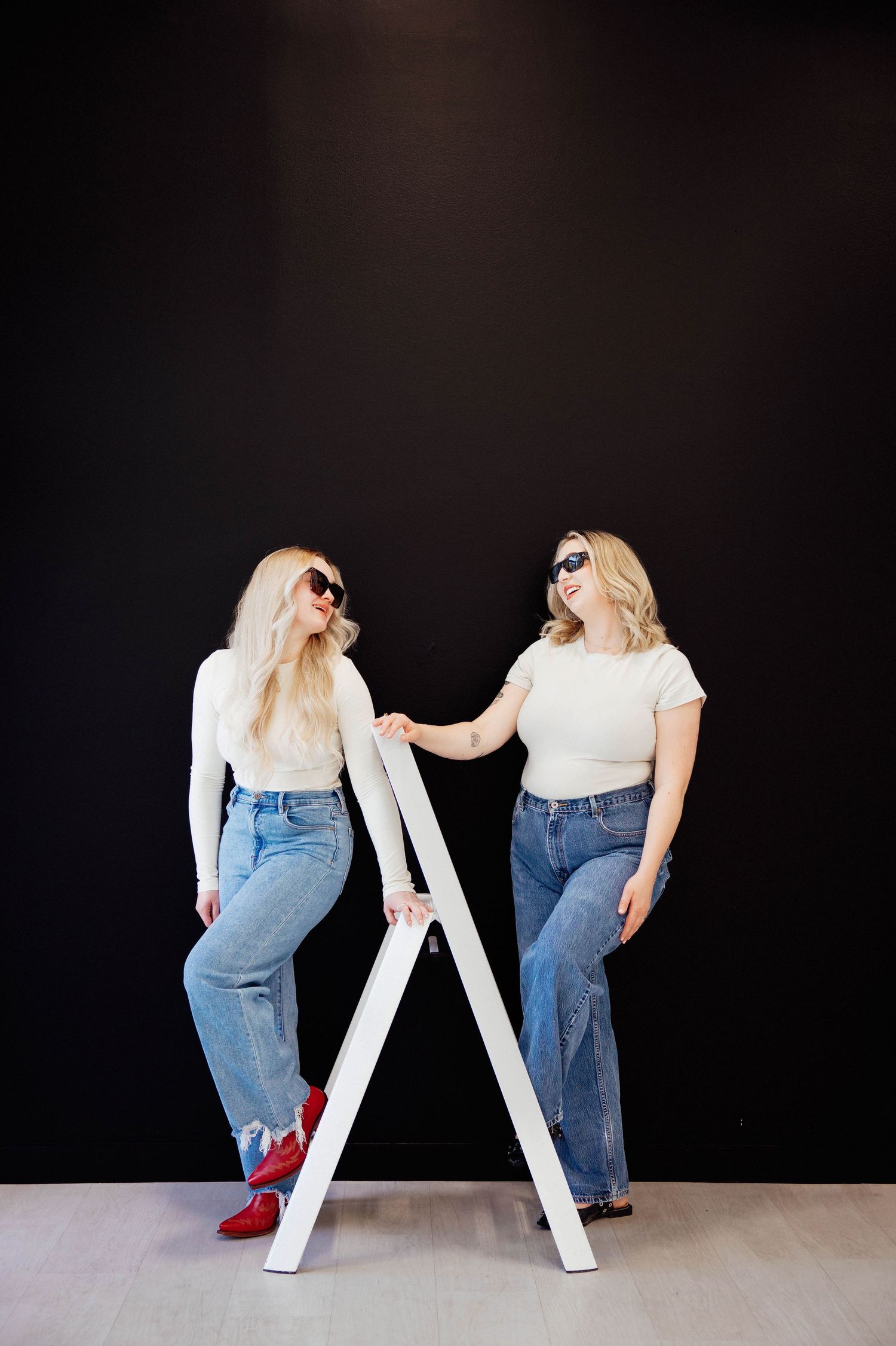 Two blonde women in white tops and jeans posing with white ladder on black wall.