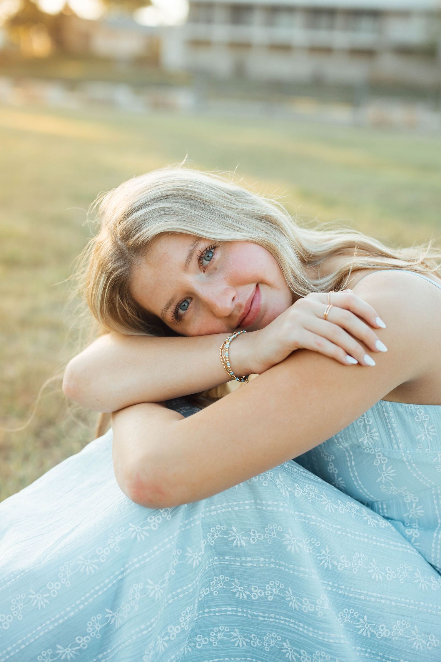 Blonde woman in light blue dress sitting in golden hour sunlight on grass, smiling softly at camera.