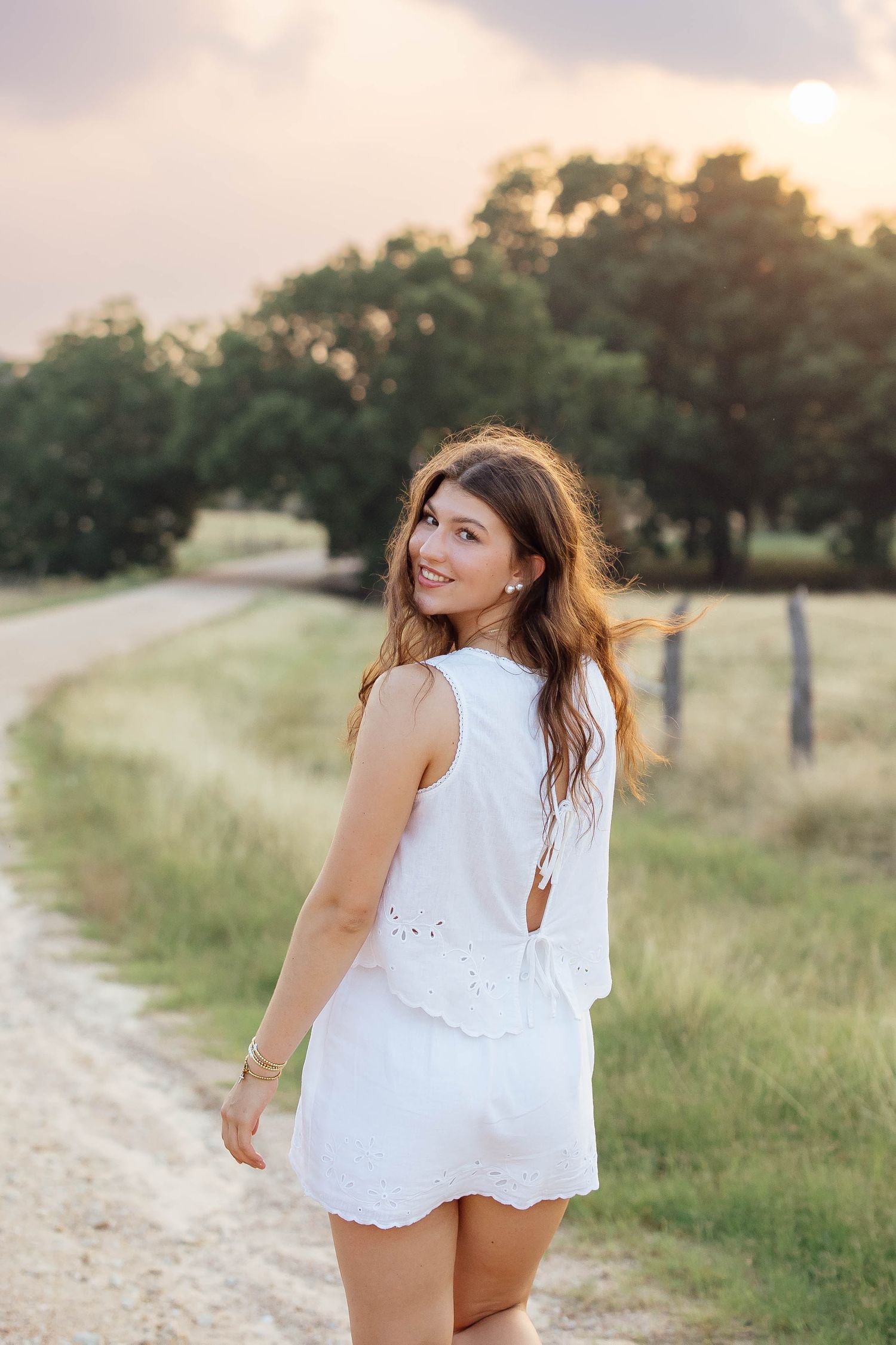 Smiling young woman in white romper on a dirt road at sunset.