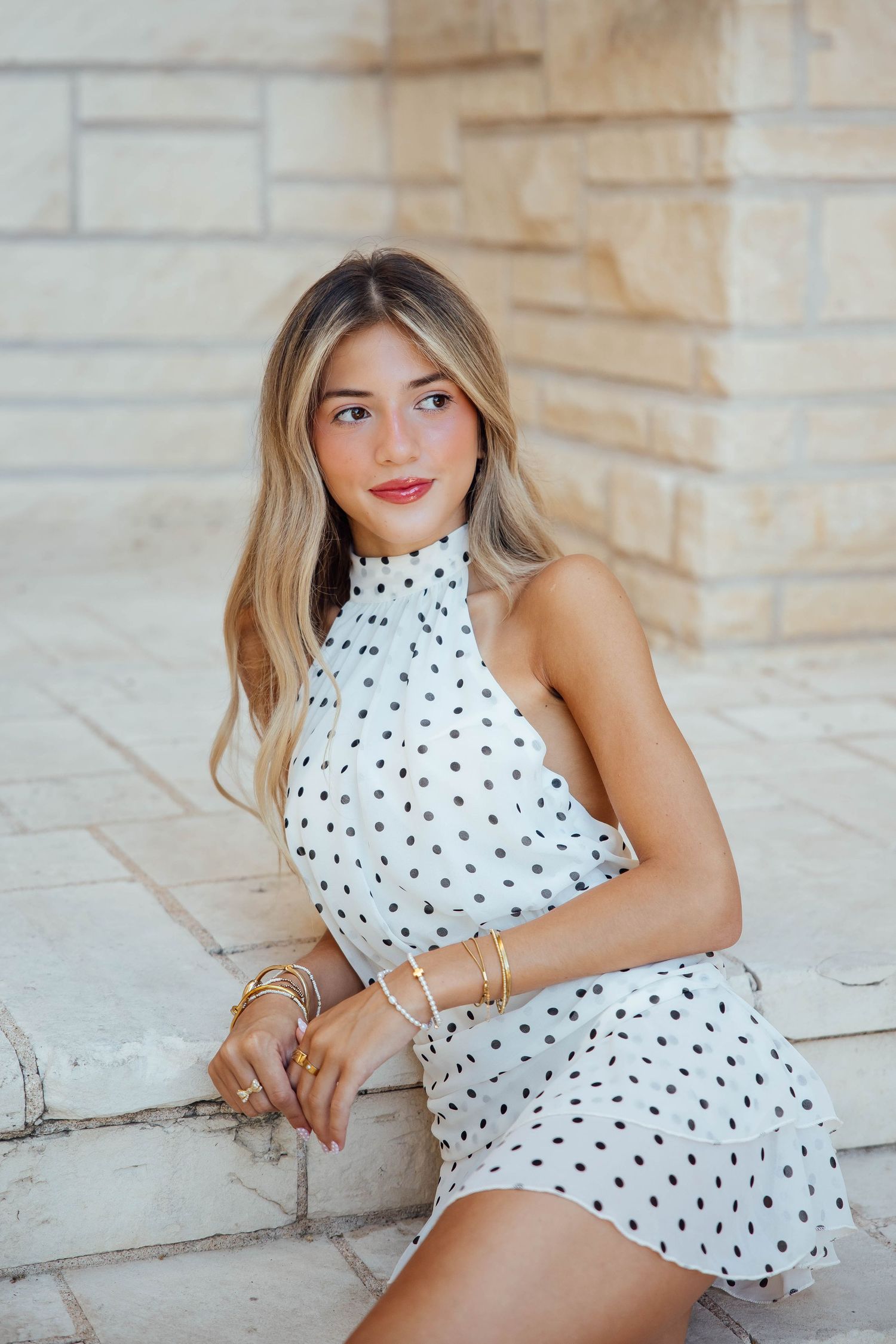 Blonde woman in white polka dot halter mini dress sitting on stone steps outdoors.