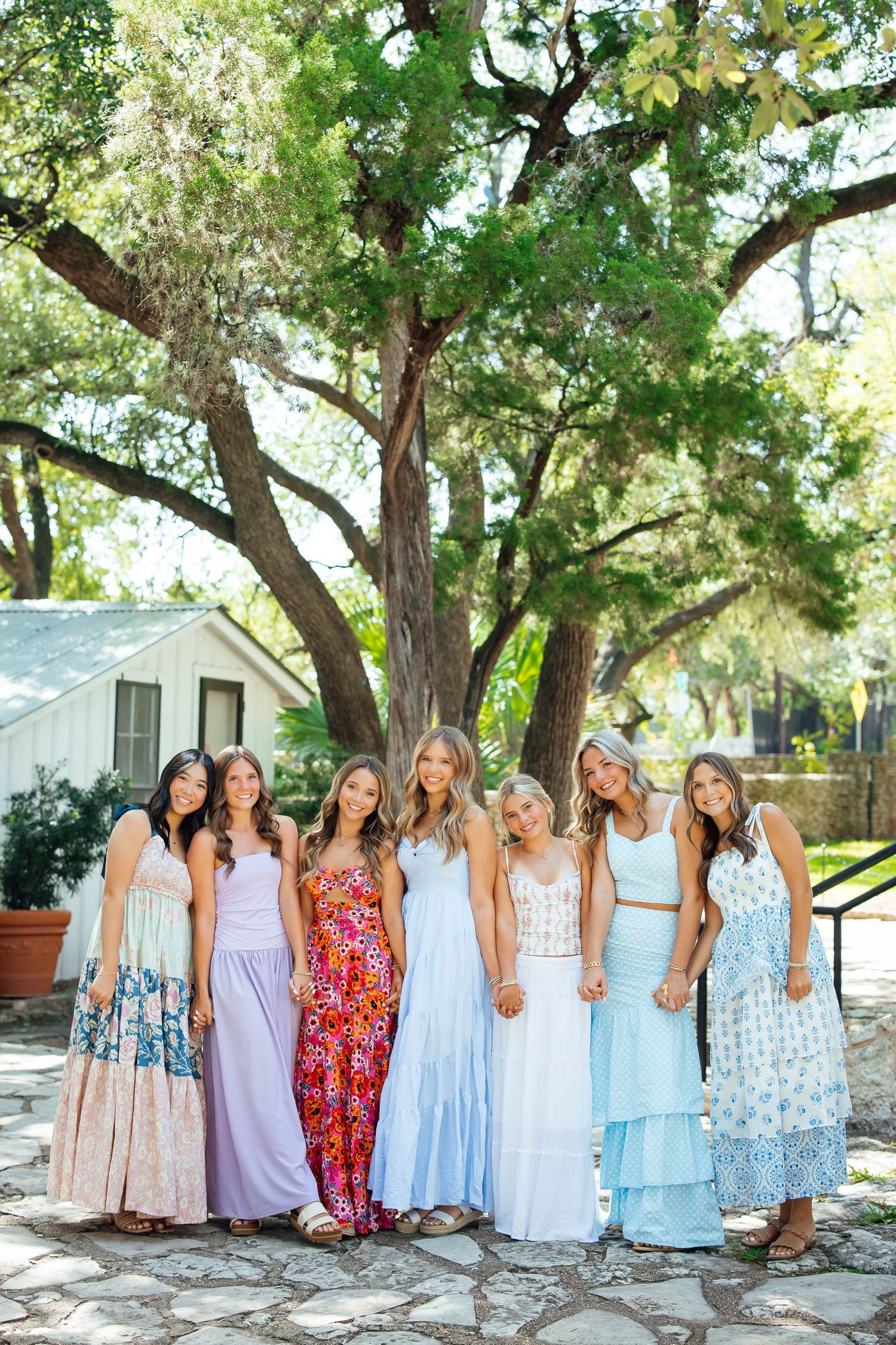 Group of seven young women in pastel maxi dresses posing under a large oak tree outdoors.