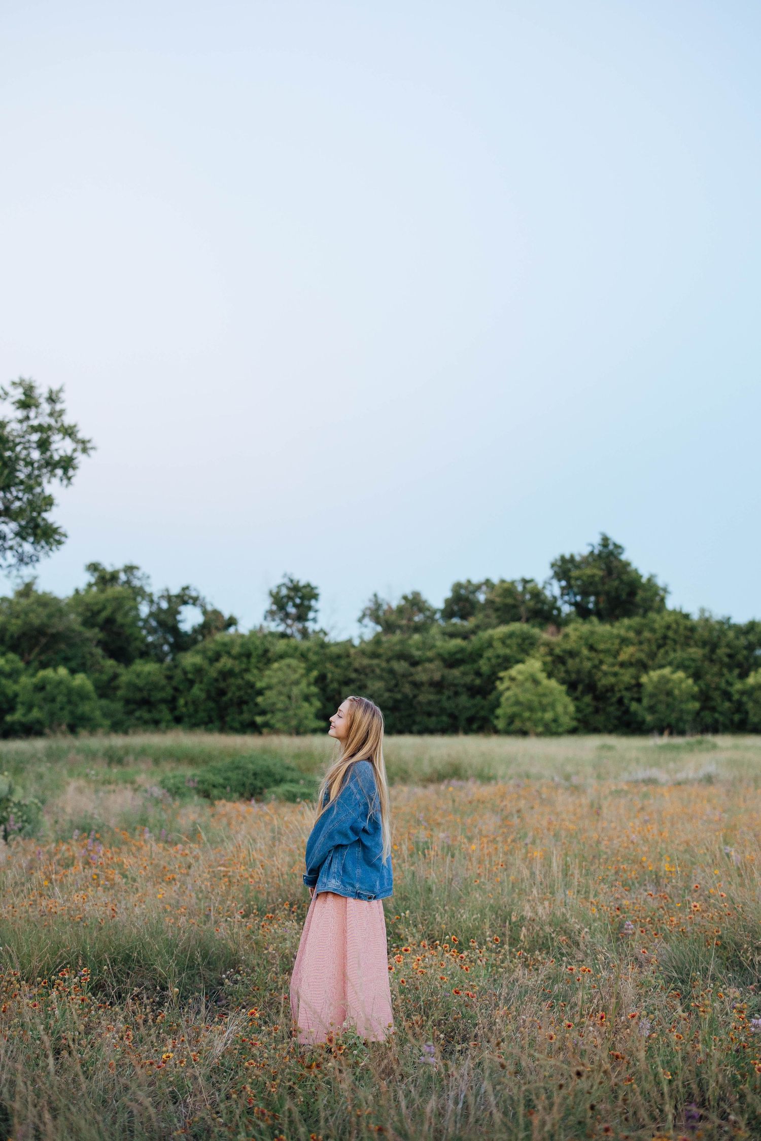 Young woman in denim jacket and pink skirt standing in wildflower meadow at dusk.