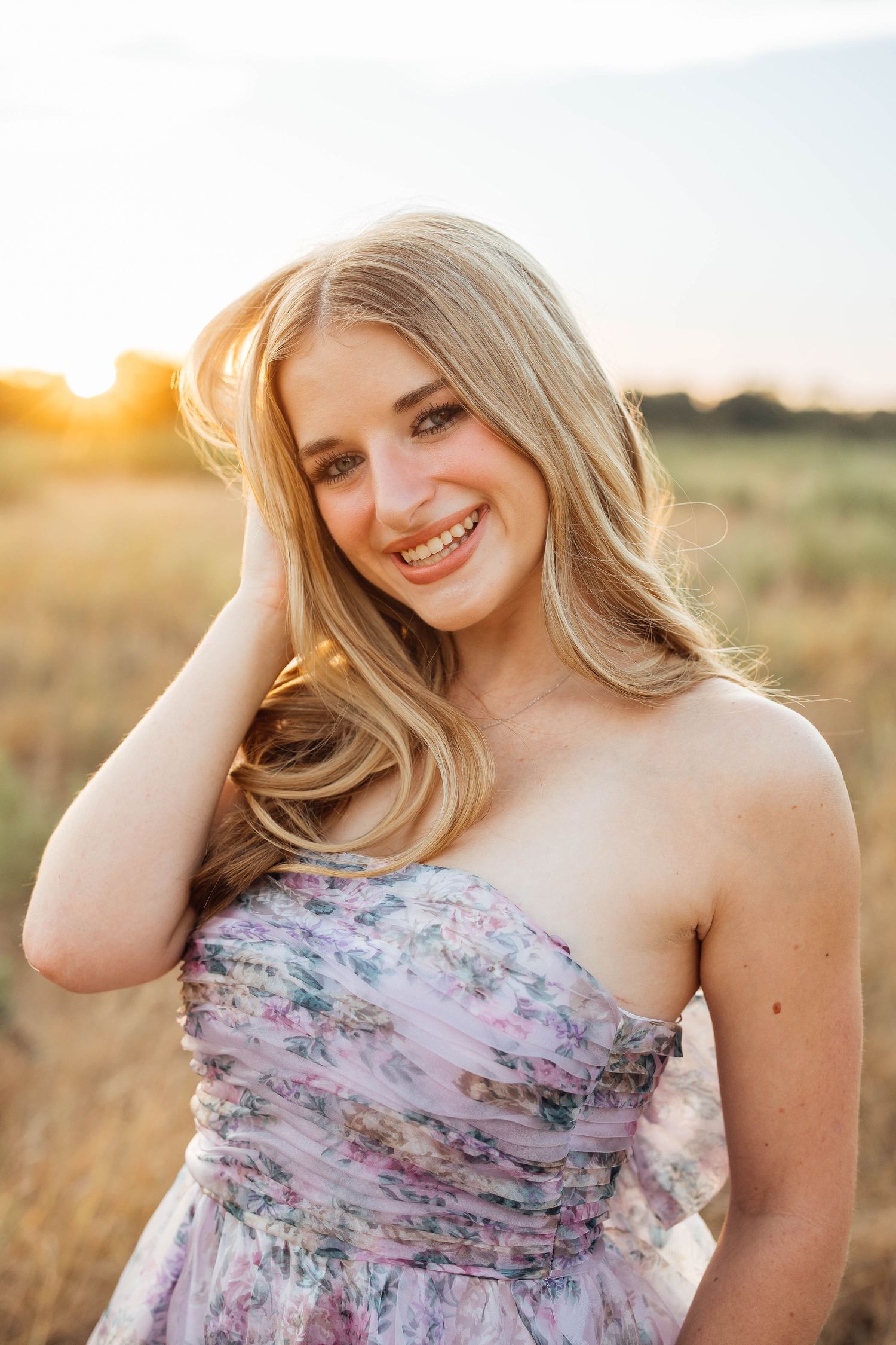 Smiling blonde teen in floral strapless dress poses in golden field during sunset for senior portrait.