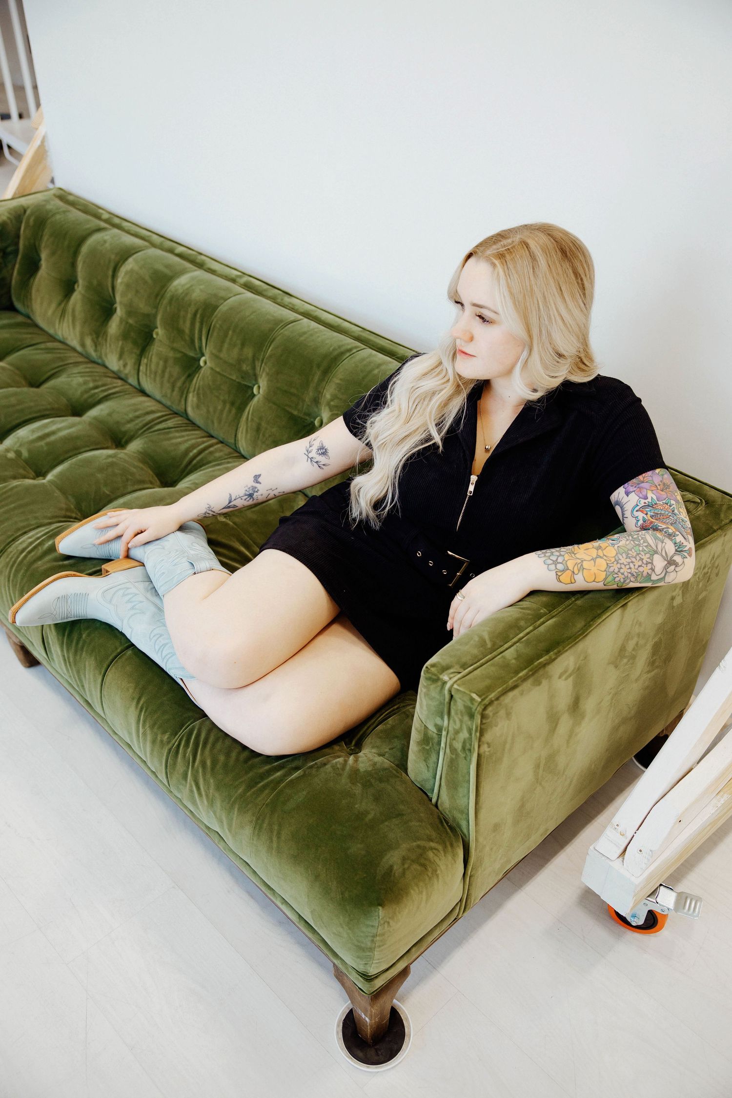 Tattooed blonde woman in black romper sitting on green velvet tufted sofa.