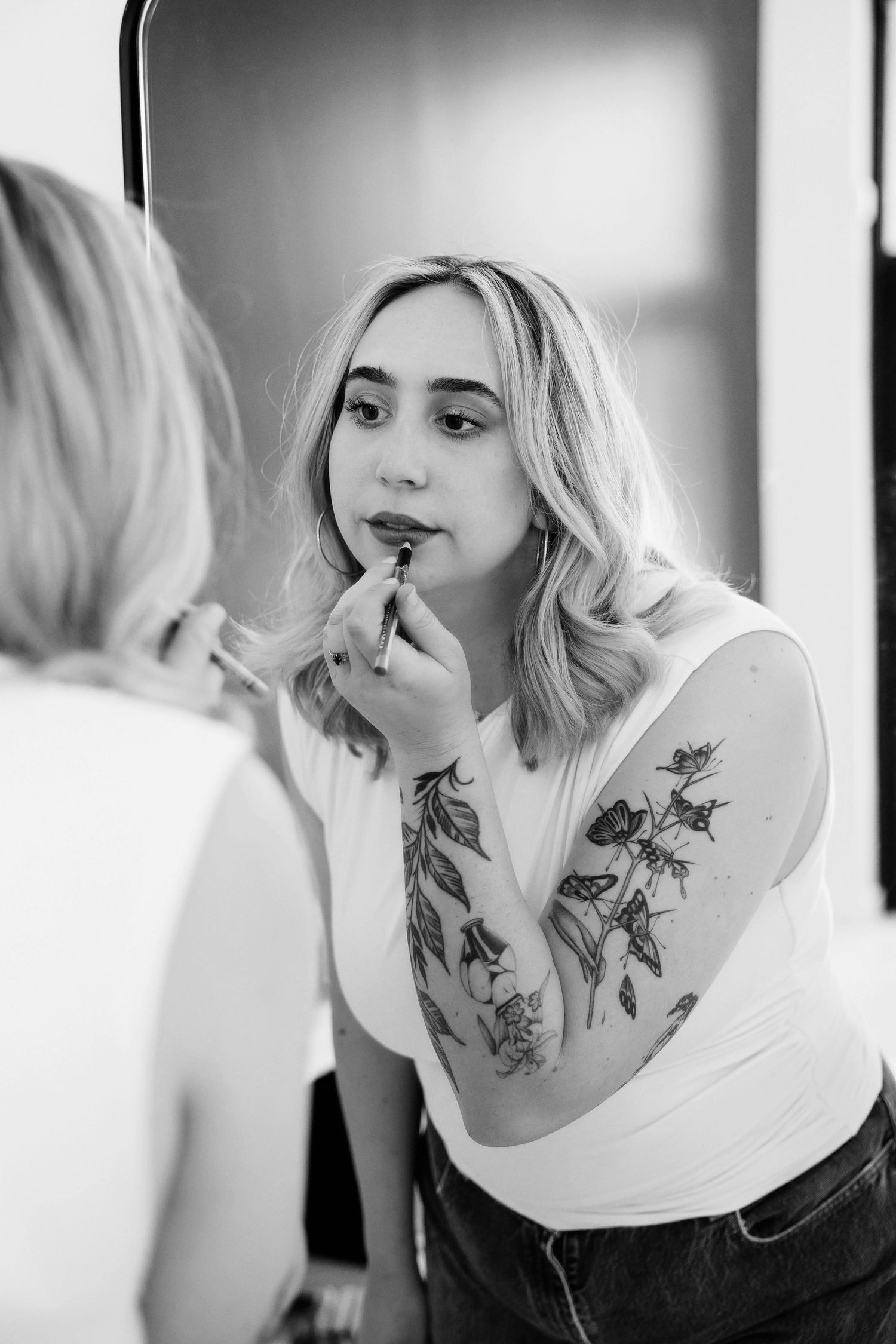 Blonde woman with floral arm tattoo applying lipstick in a mirror, black and white photo.