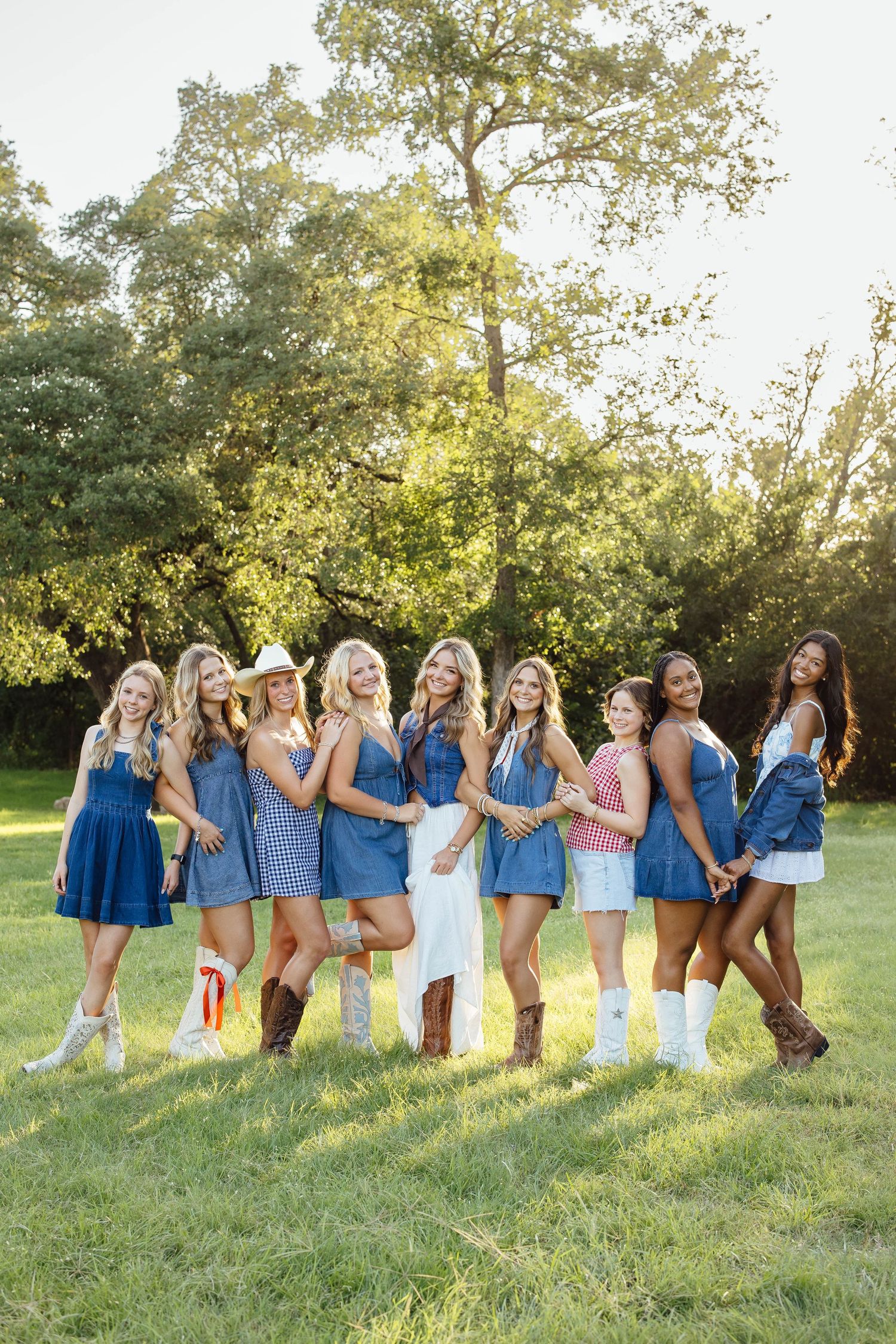 Group of young women in denim outfits and cowboy boots posing together outdoors in a sunny green field.