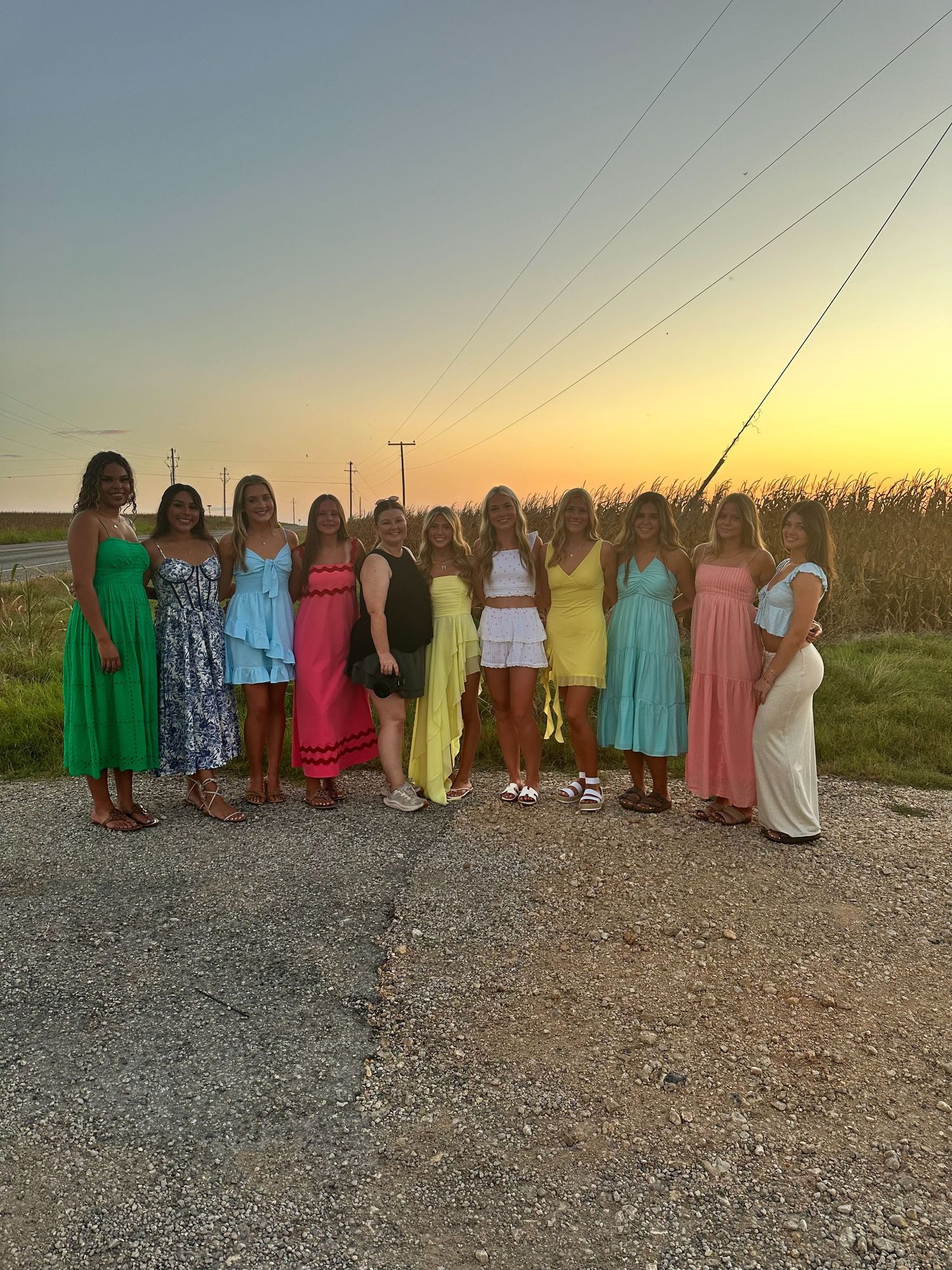 Group of women in colorful summer dresses posing together at sunset on a rural road.