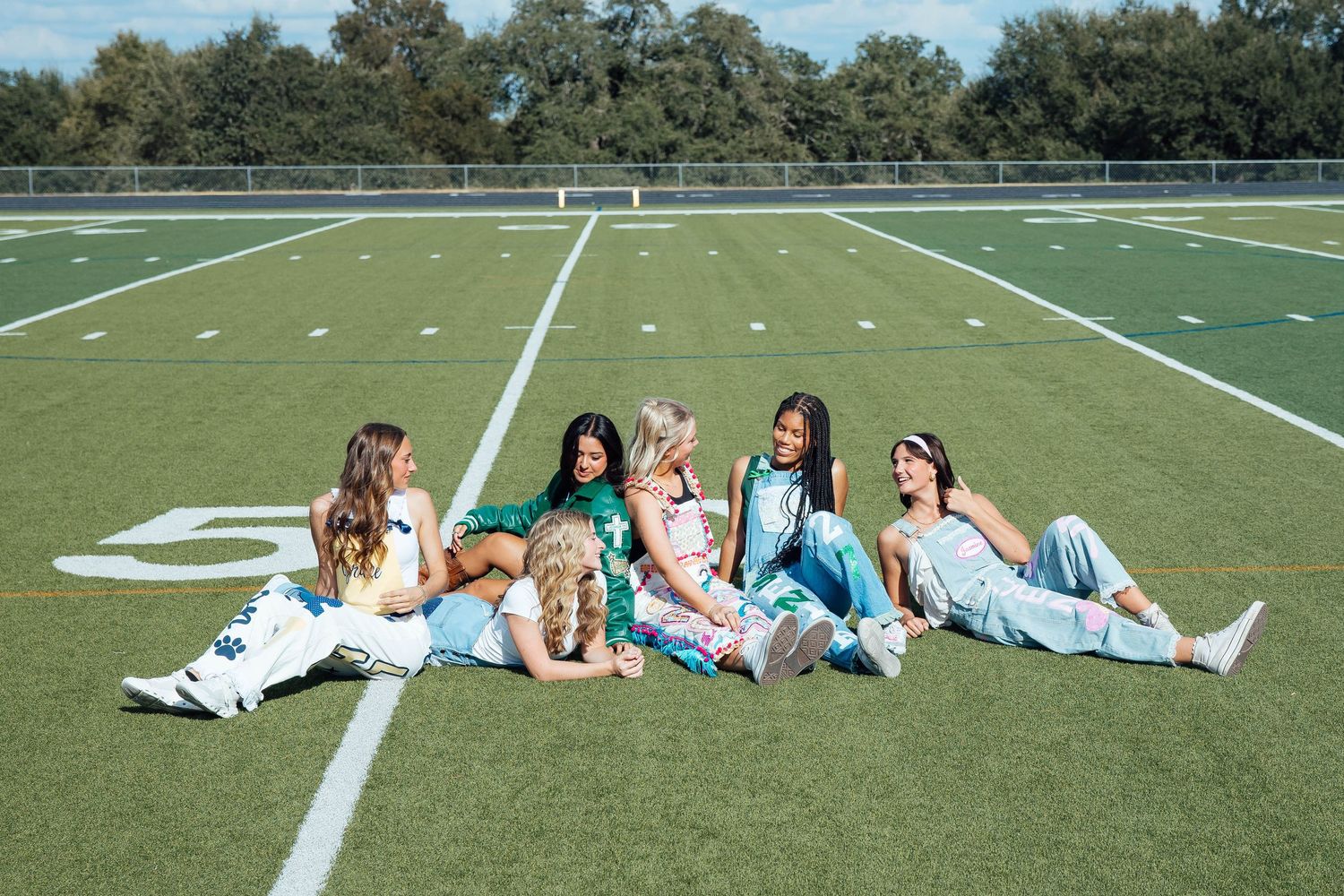 Six young women in tie-dye outfits sit together on a football field turf.