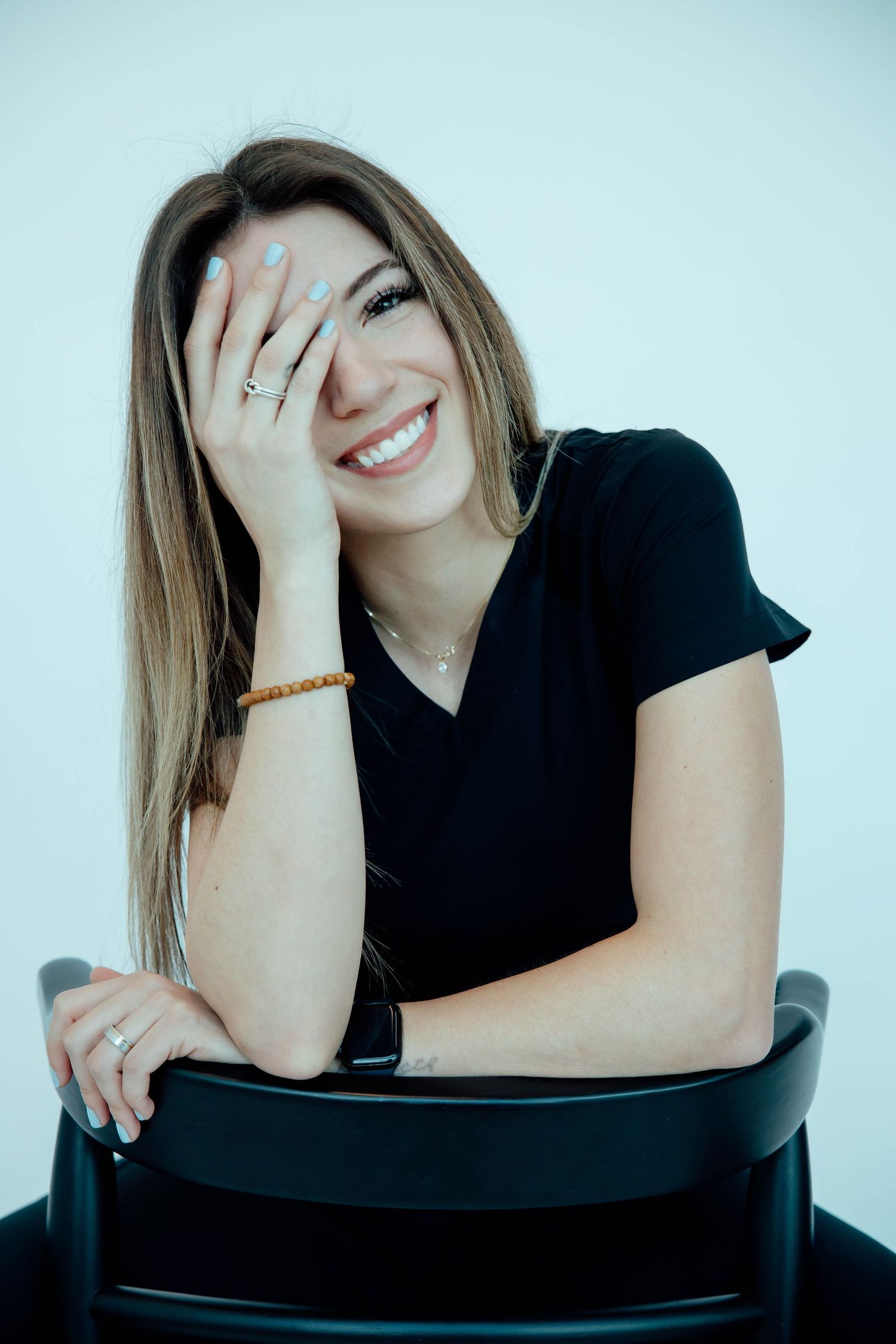 Smiling woman with long hair sitting on a black chair, resting her hand on her face.