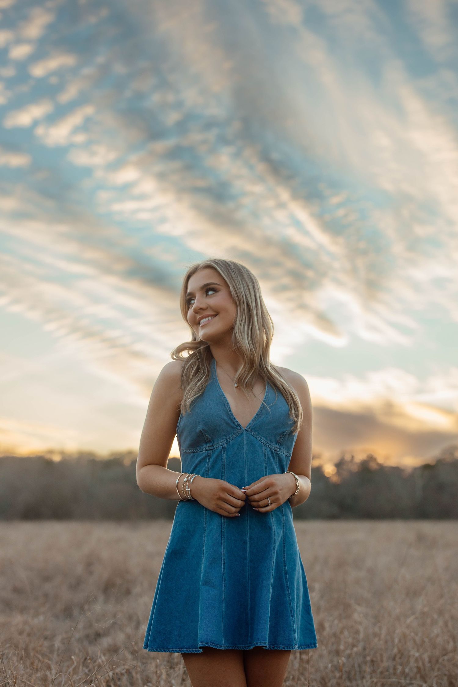 Blonde young woman in blue mini dress smiling in a golden field under a dramatic sunset sky.