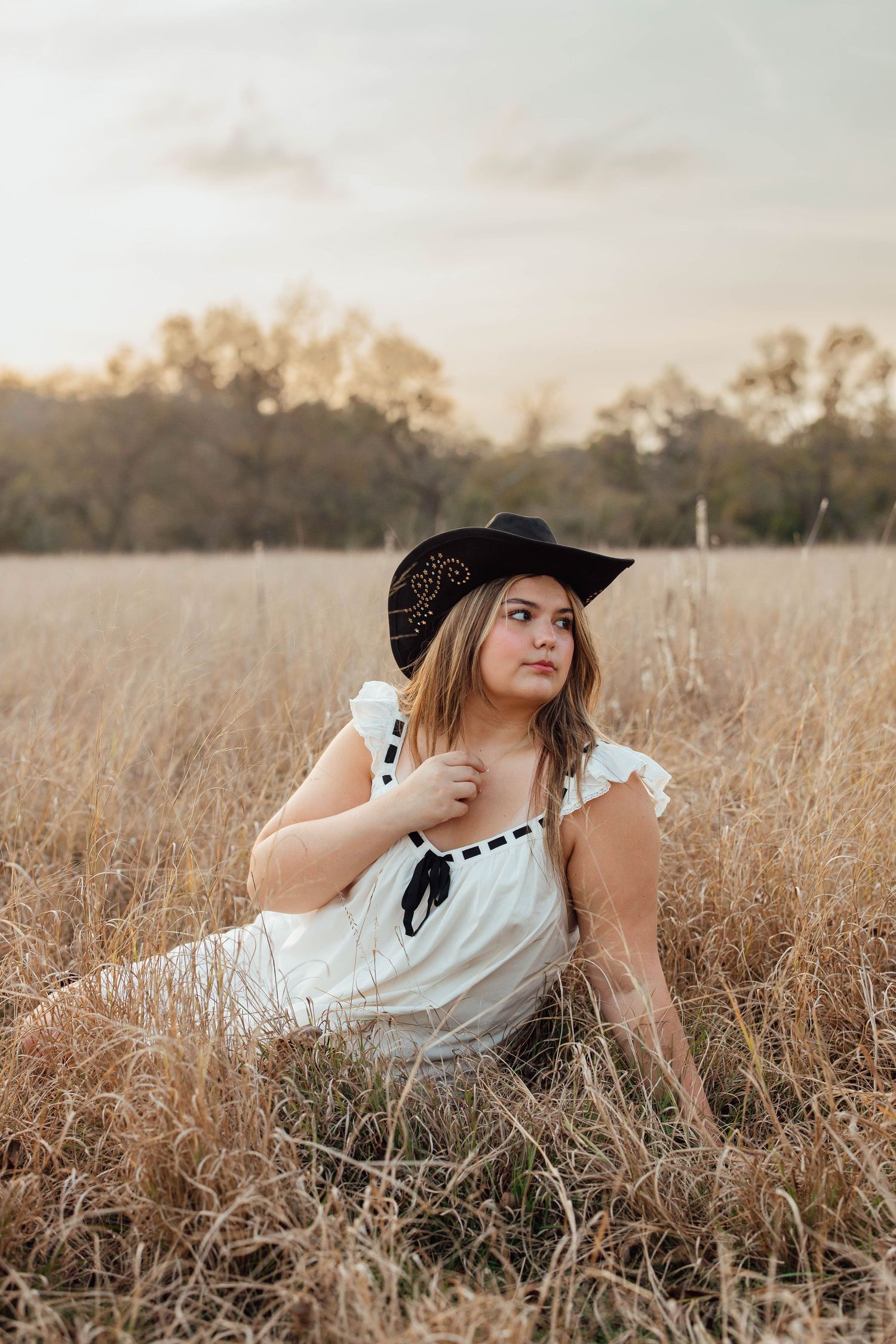 A young woman in a white dress and black cowboy hat sits in a golden field at sunset.