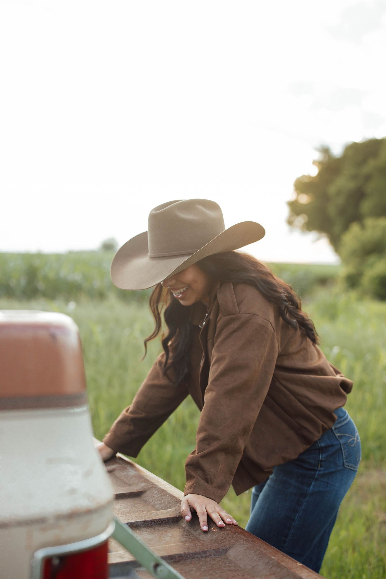 Smiling woman in cowboy hat and brown jacket leaning on vintage truck tailgate in rural field.
