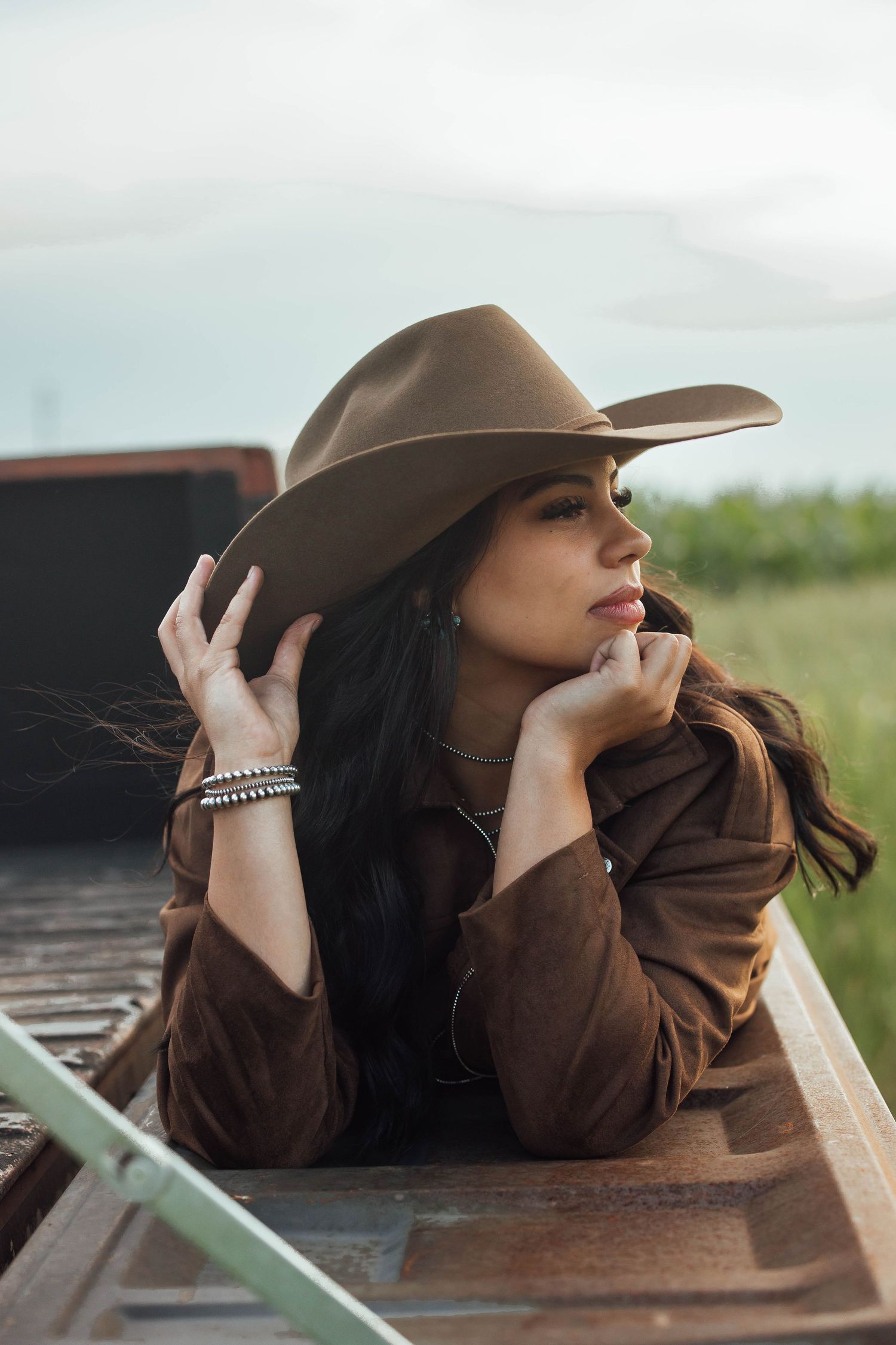 A brunette woman in a brown suede jacket adjusts her tan cowboy hat while leaning on a truck bed.