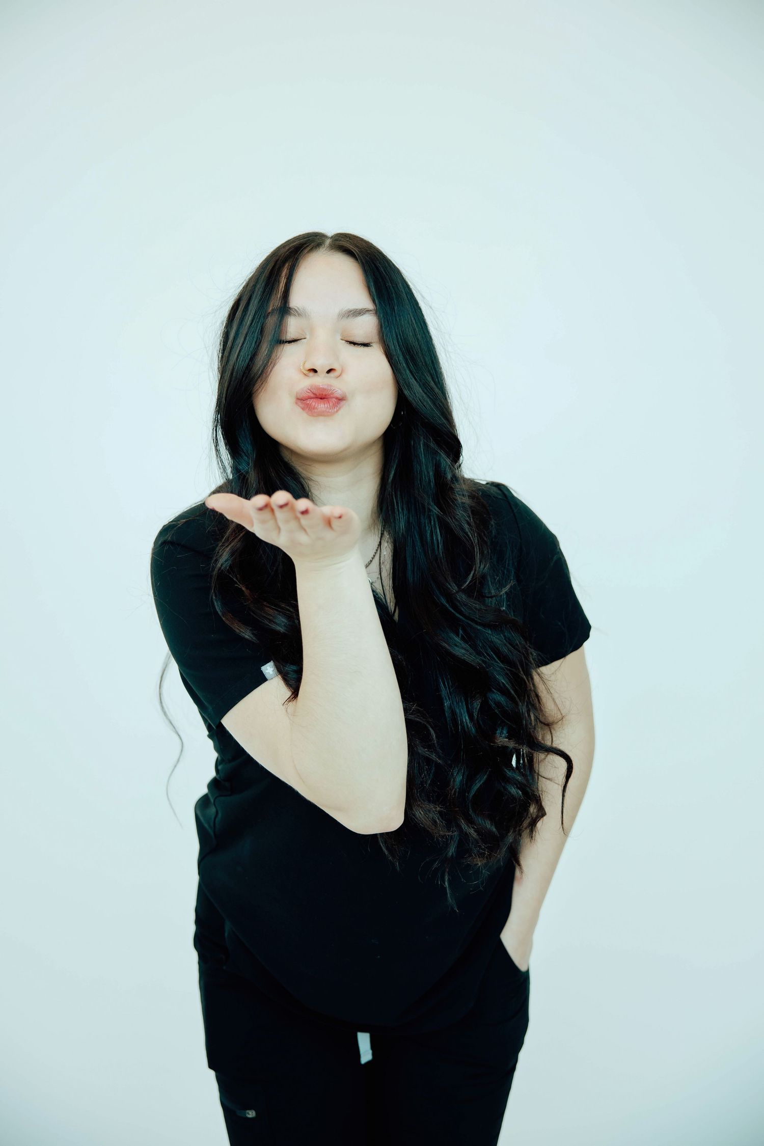 Young woman with long dark wavy hair blowing a kiss against a light background.