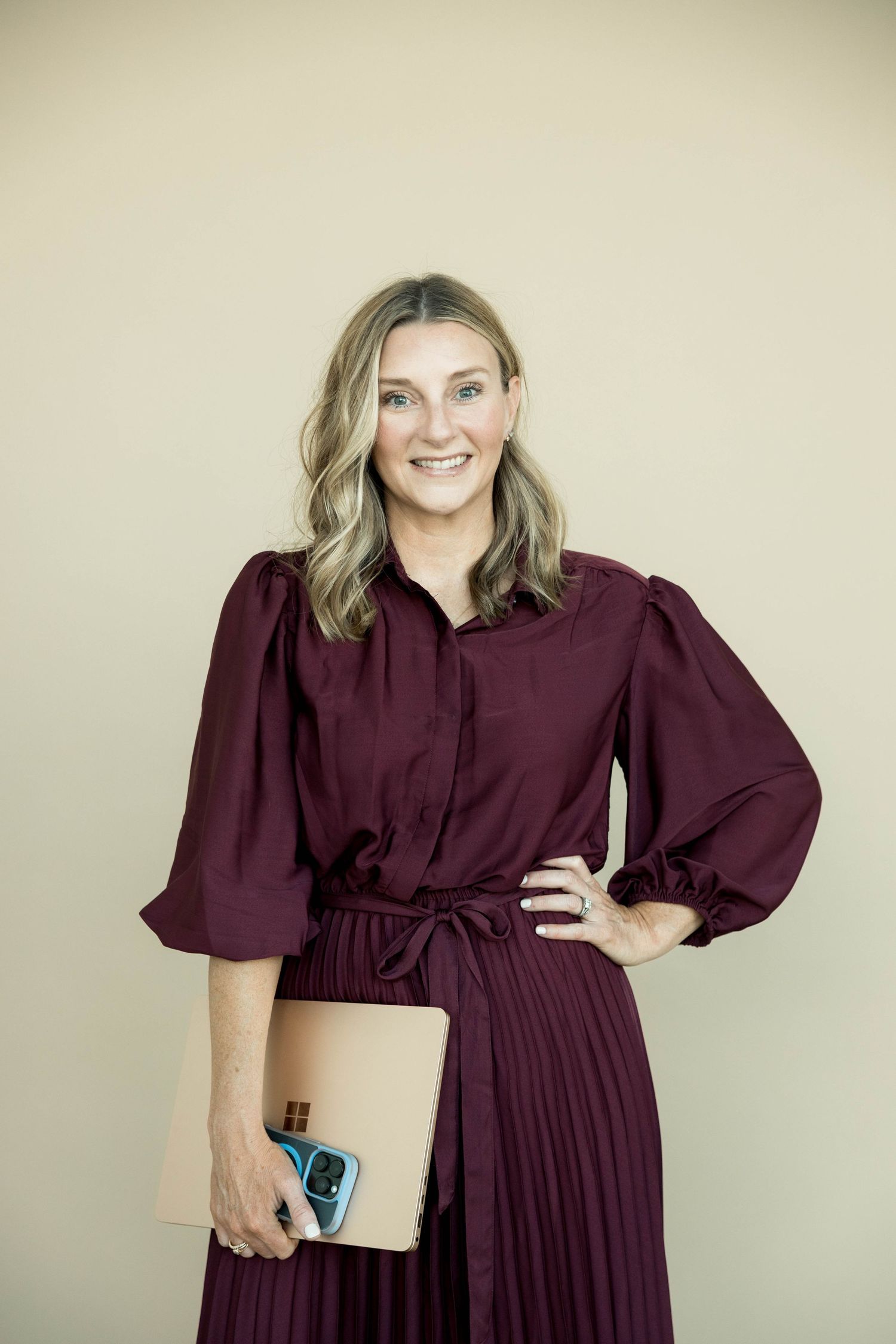 Smiling blonde woman in burgundy pleated dress holding a notebook and phone against a beige wall.