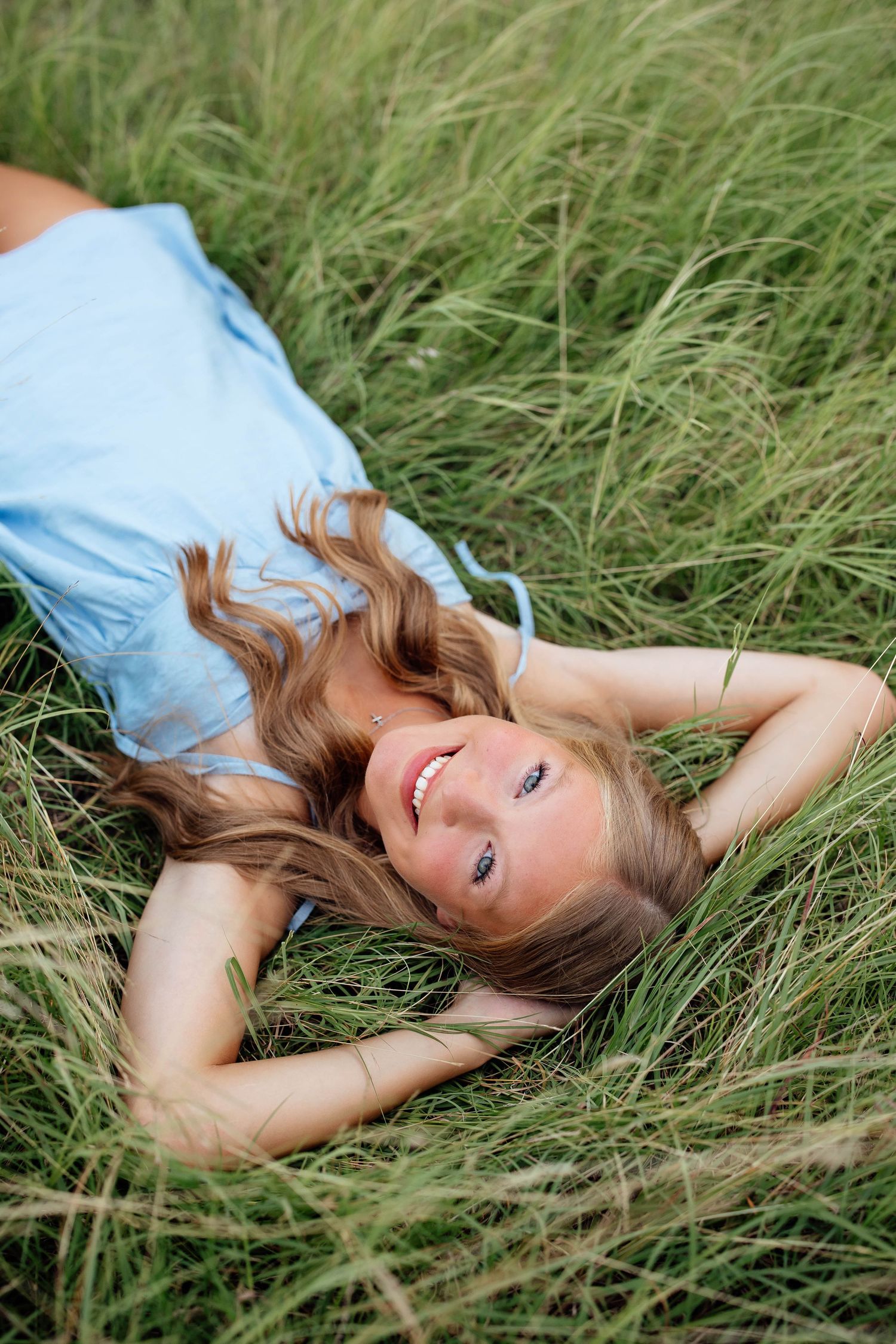 Smiling teenage girl with long hair lying in tall green grass wearing a light blue dress.