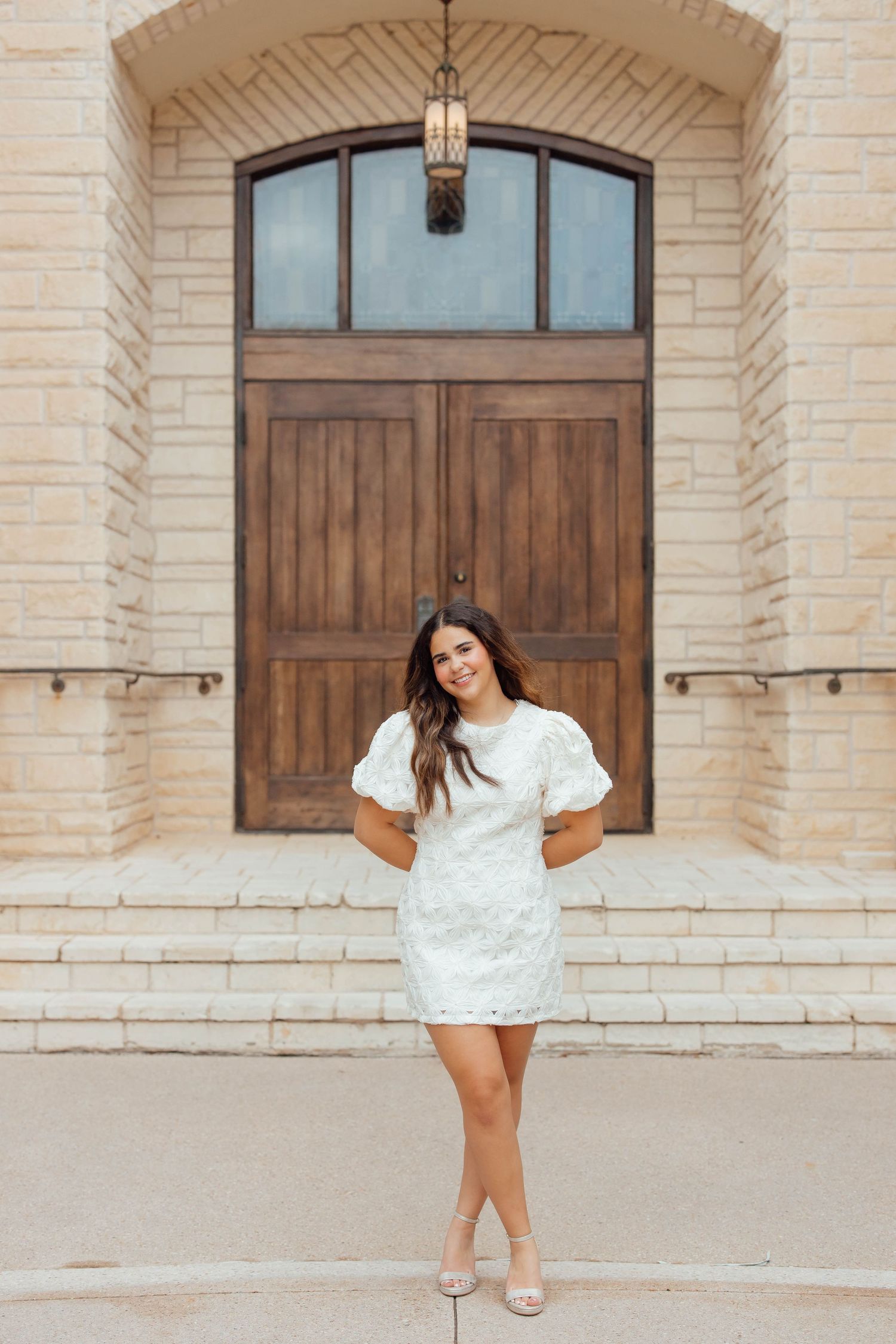 Smiling brunette in white lace puff-sleeve dress standing before elegant wooden double doors.