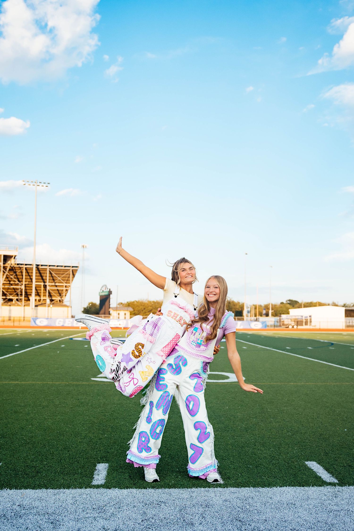 Two girls posing on football field, one wearing colorful patterned pants, under blue sky.