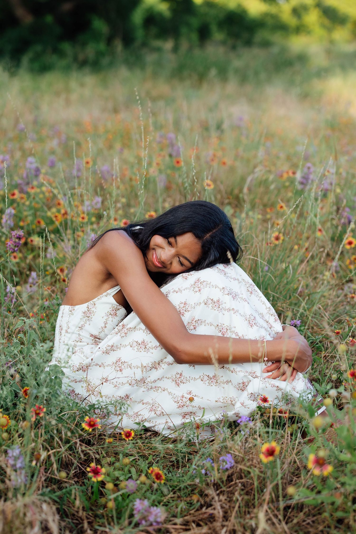 Young woman in floral skirt sitting in a wildflower meadow hugging a decorative pillow.