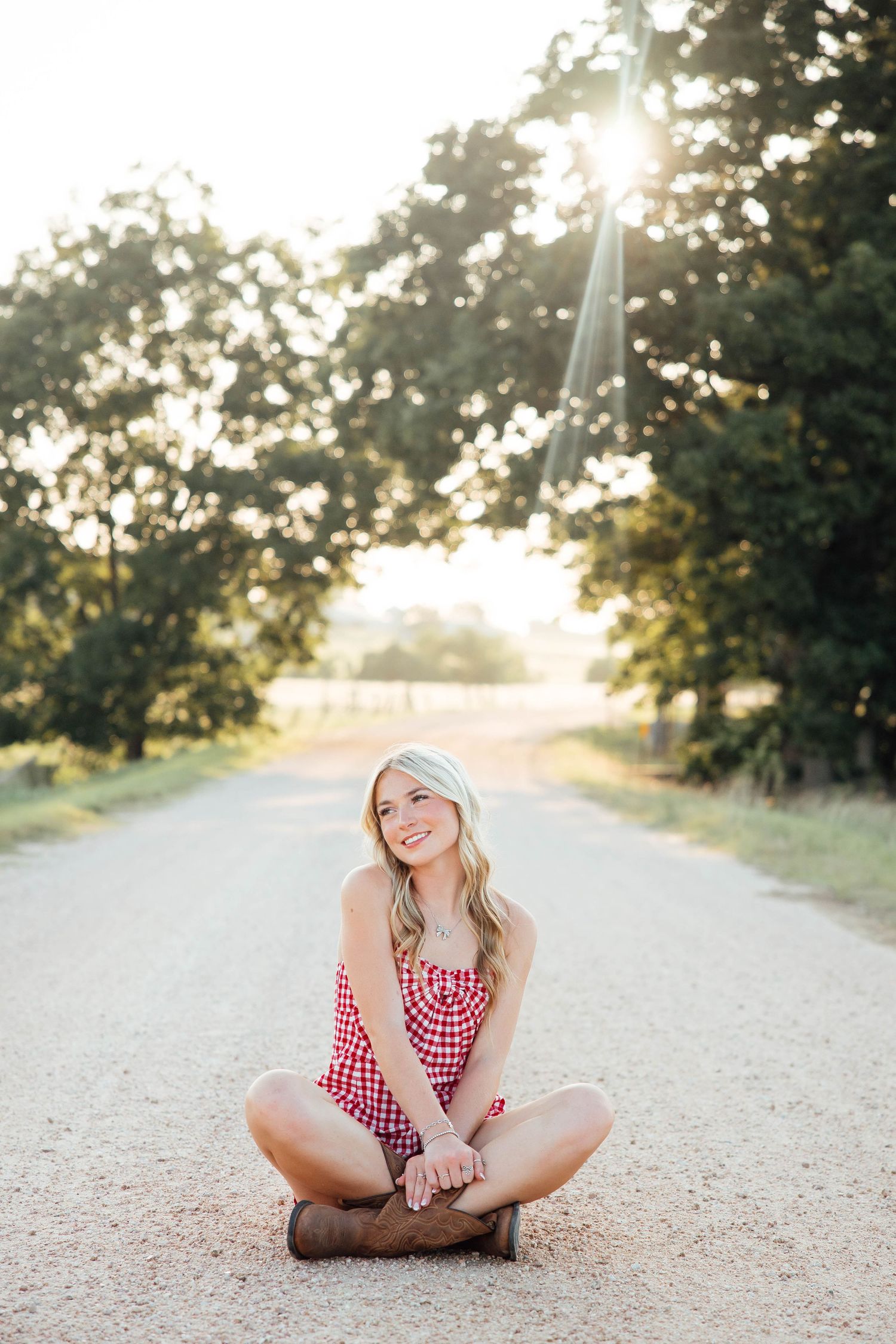 Smiling blonde girl in red gingham romper sits cross-legged on a sunny tree-lined dirt road.