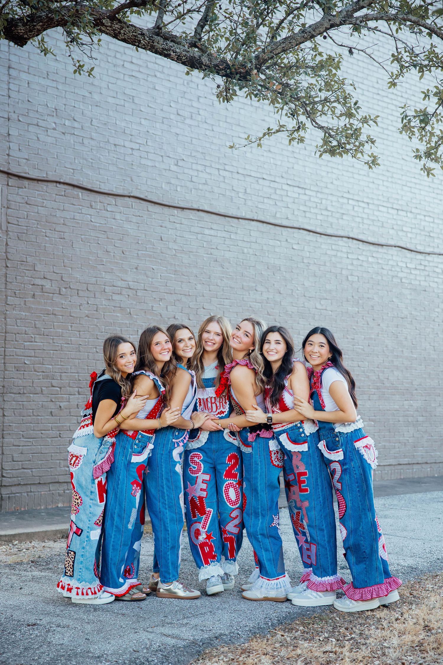 Group of seven teen girls posing in decorated senior jeans against a brick wall.