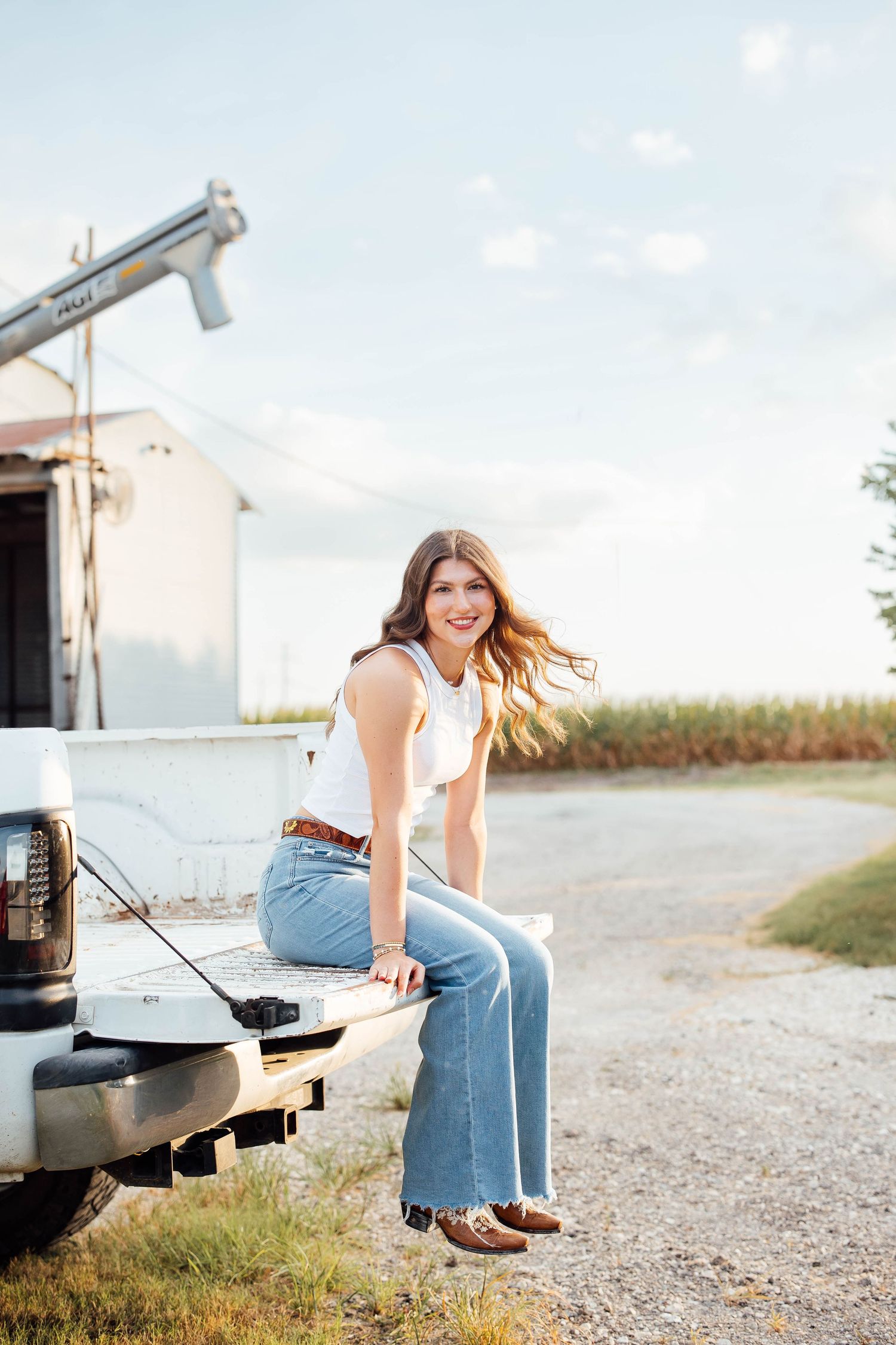 Smiling brunette girl in white tank and flare jeans sitting on truck tailgate.