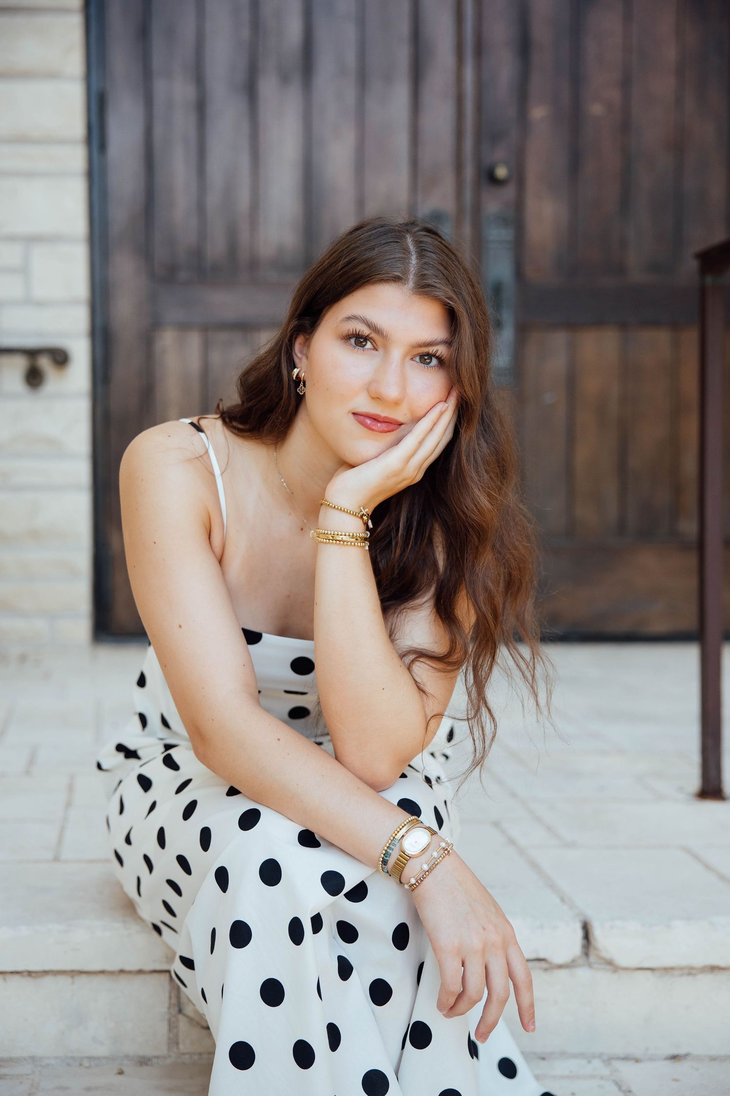 Brunette woman in white polka dot dress sitting on steps near a rustic wooden door.