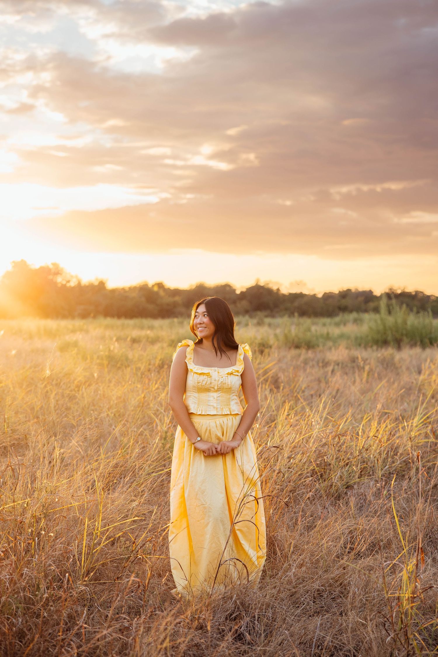 Young woman in yellow maxi dress stands in golden field during dramatic sunset senior photo.