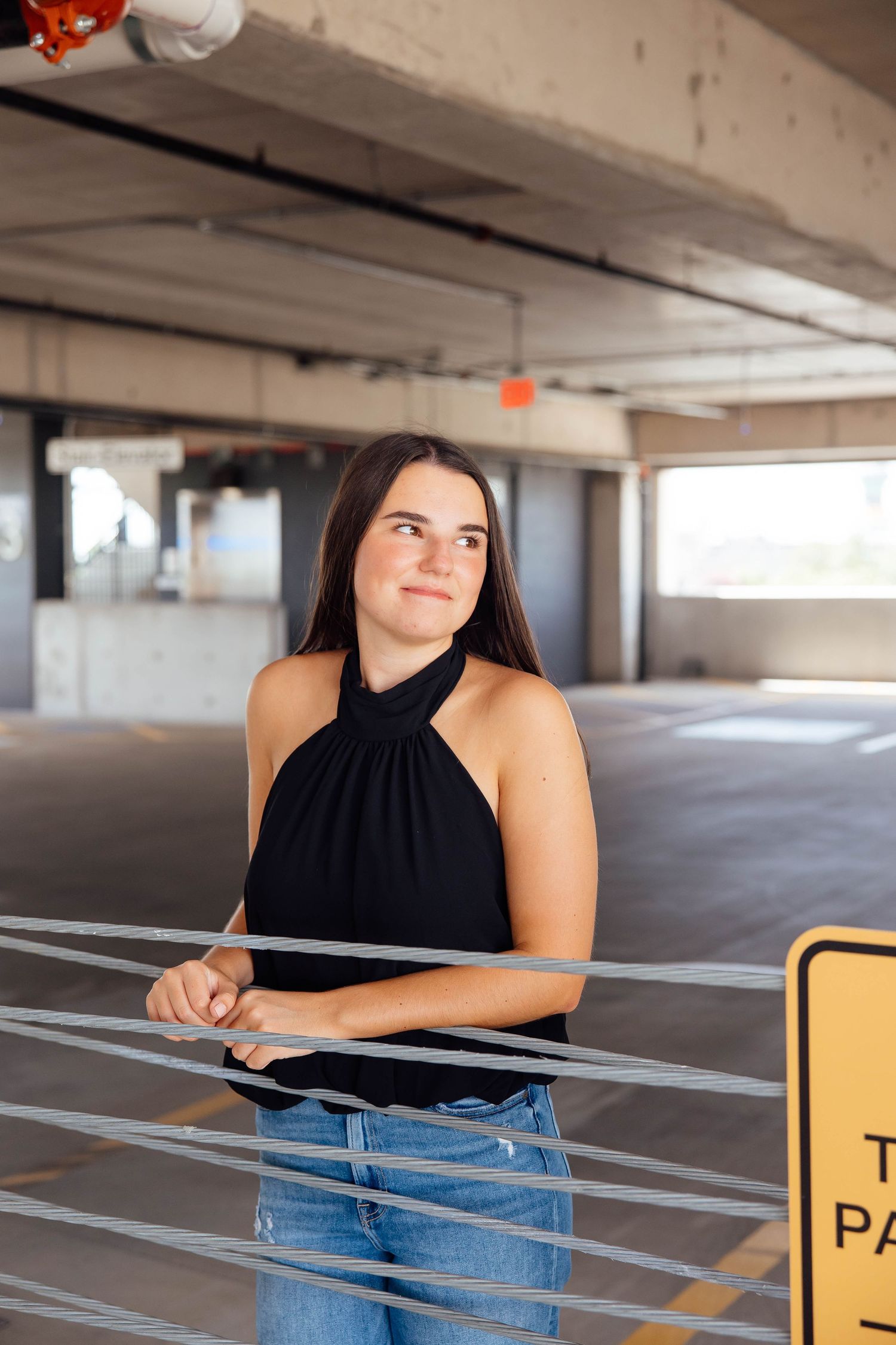 Young woman in black halter top and jeans poses in urban parking garage with cable barriers.