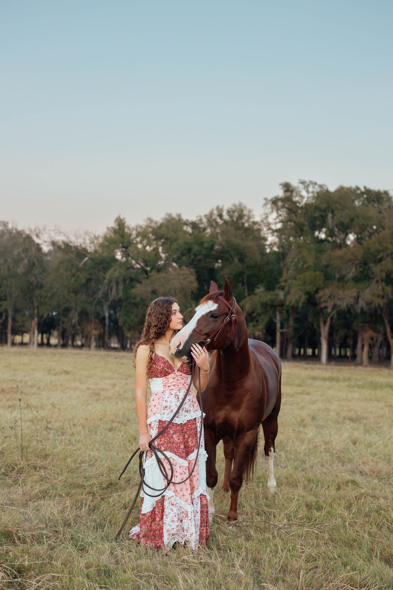 Young woman in floral maxi dress affectionately petting a brown horse in a green pasture.